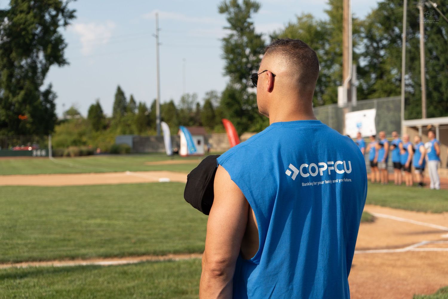 Man in blue COPFCU sleeveless shirt watches players on a sunny outdoor baseball field.