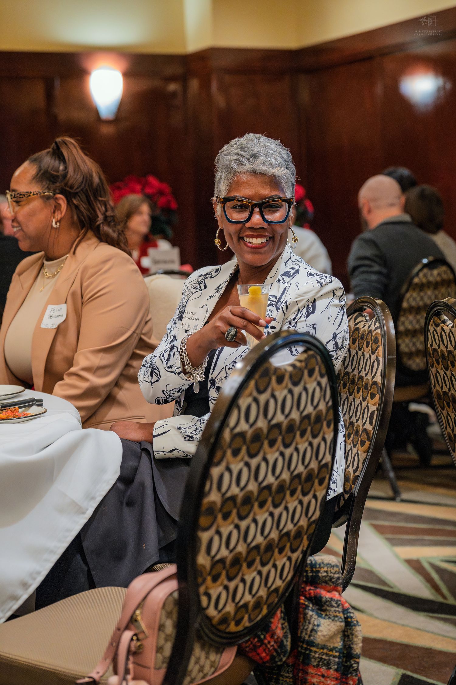 Smiling woman holding a drink at an elegant indoor event, seated among guests in a formal dining room.