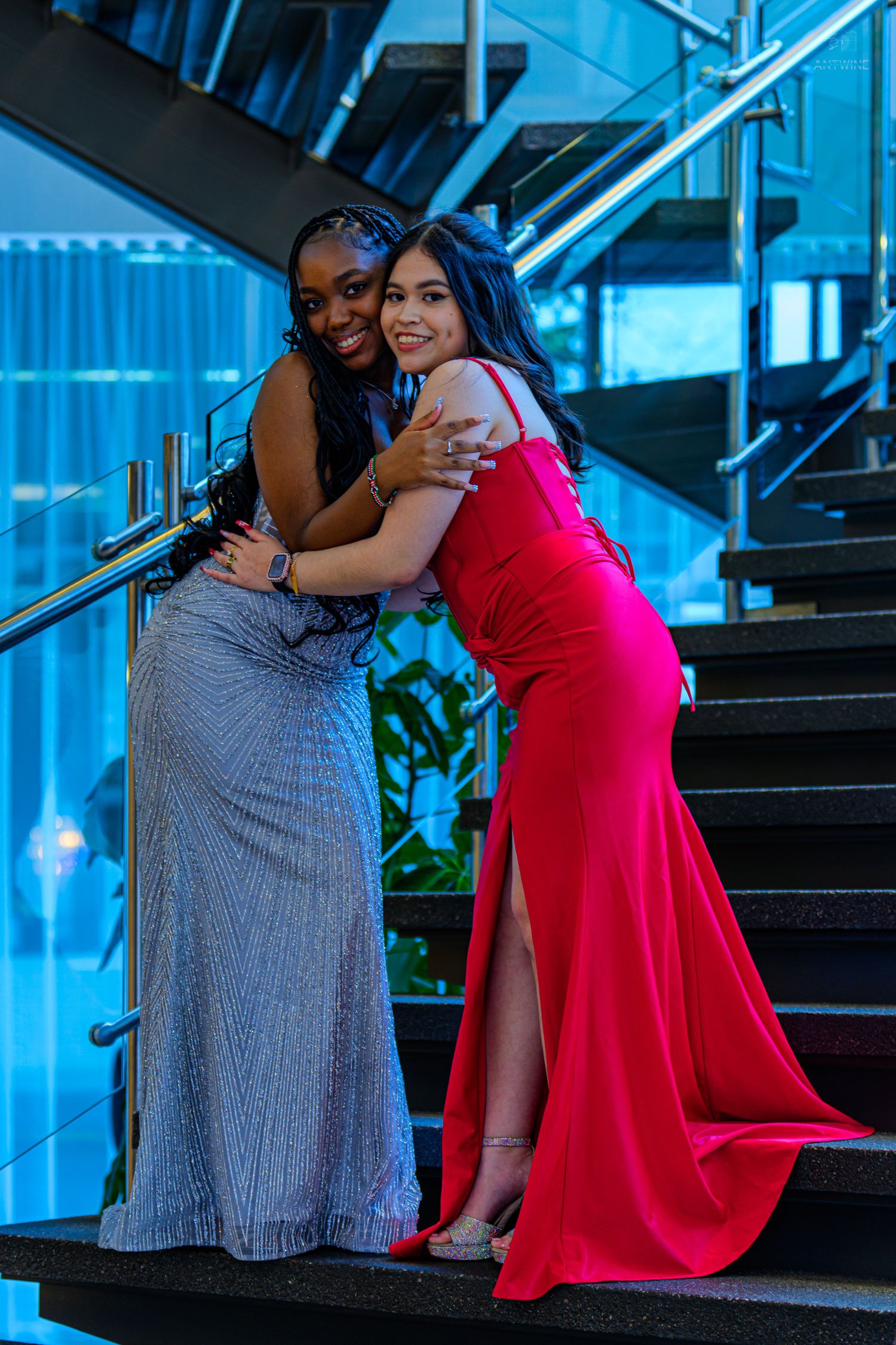 Two young women in elegant formal gowns, one silver and one red, hugging on stairs at a prom event.