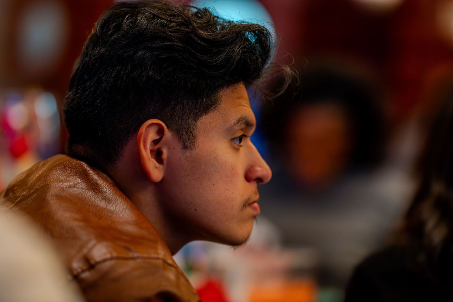 Young man in brown leather jacket listening attentively in a crowded indoor setting, captured in profile view.