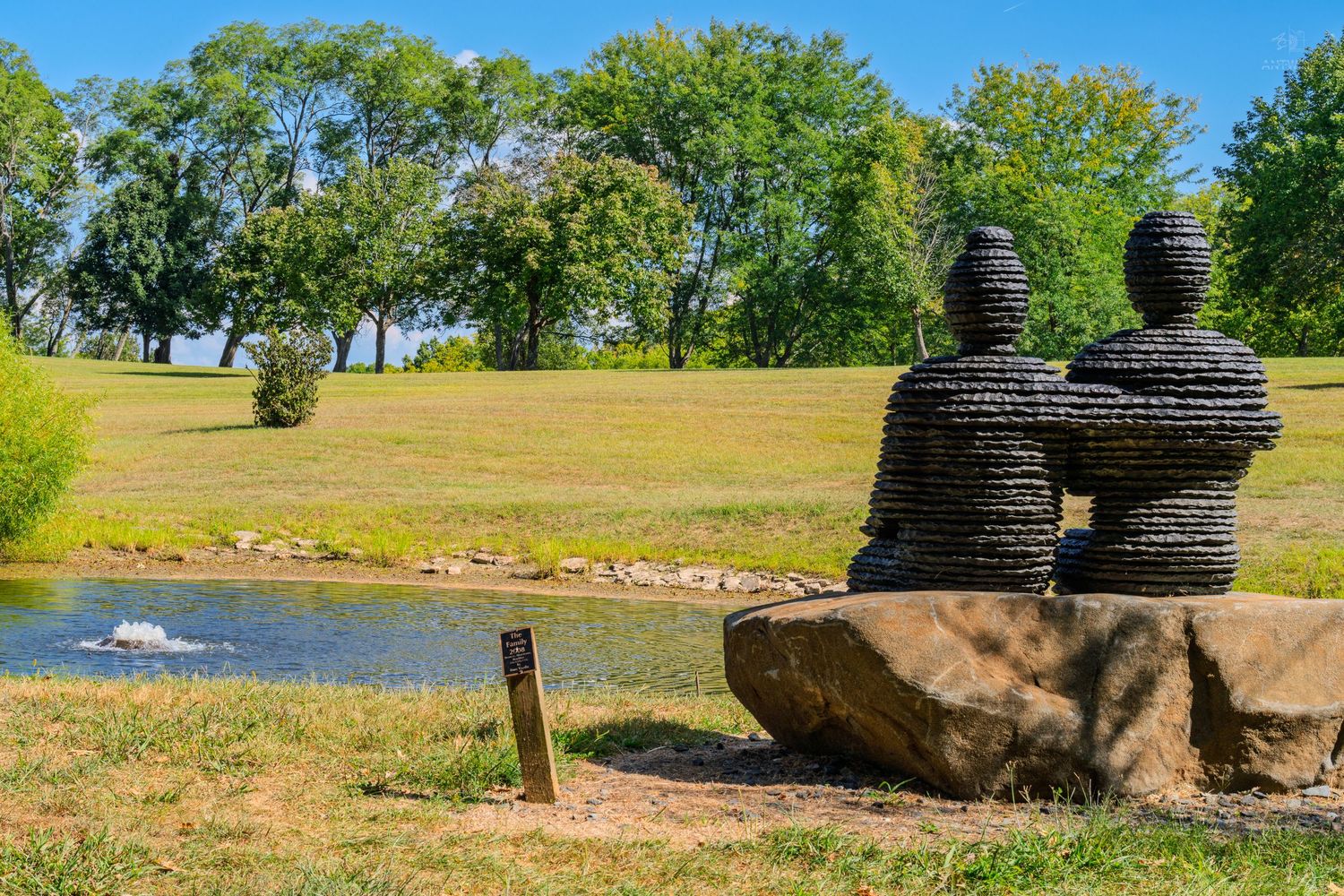 Two wire sculptures of figures sitting on a rock beside a tranquil pond in a lush green park.