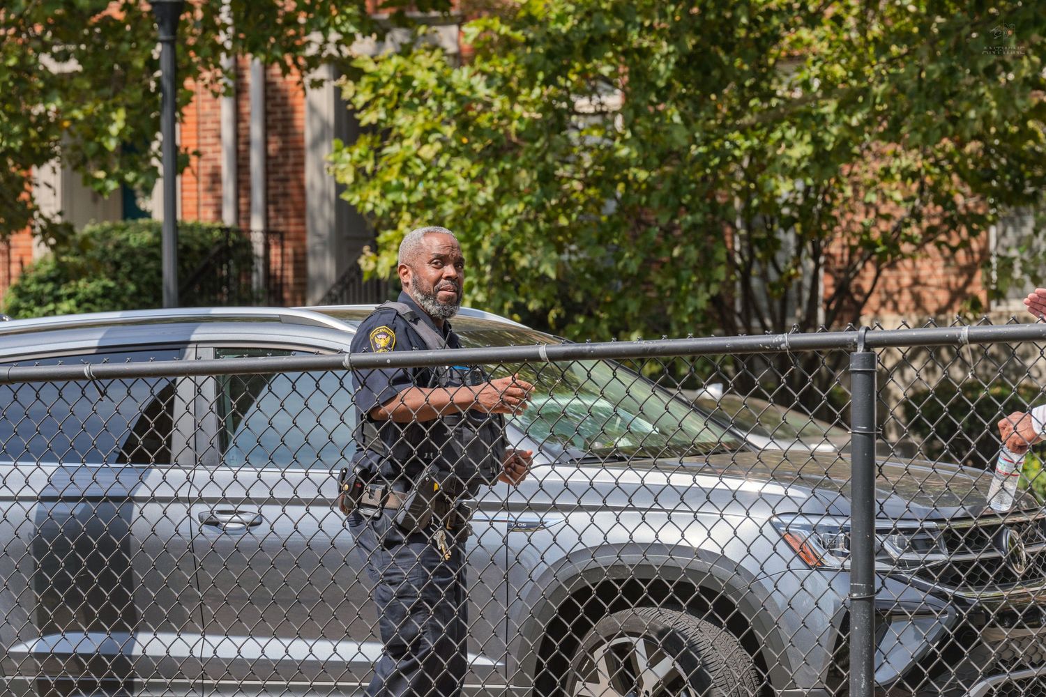 A police officer stands beside a chain-link fence near parked cars on a city street.