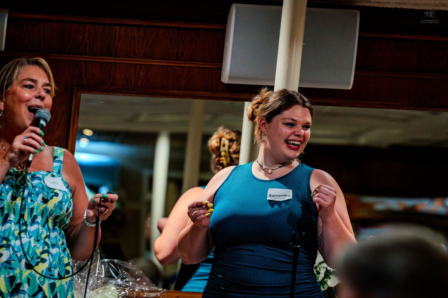 Two women at an event, one speaking into a microphone while the other celebrates excitedly.