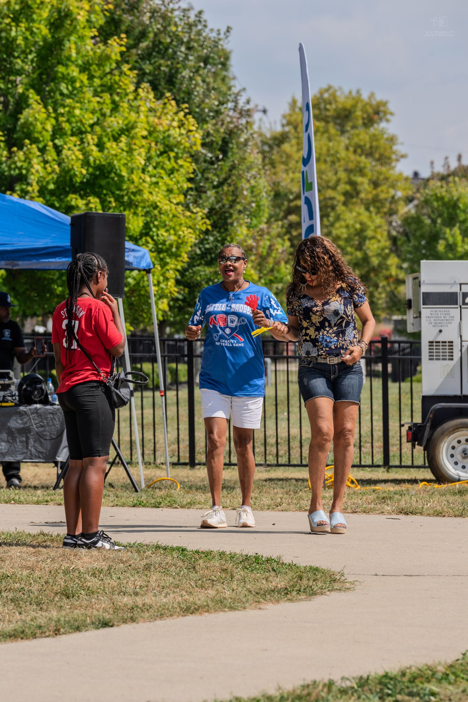Three people conversing on a sunny sidewalk at an outdoor community event with blue tent and banner.