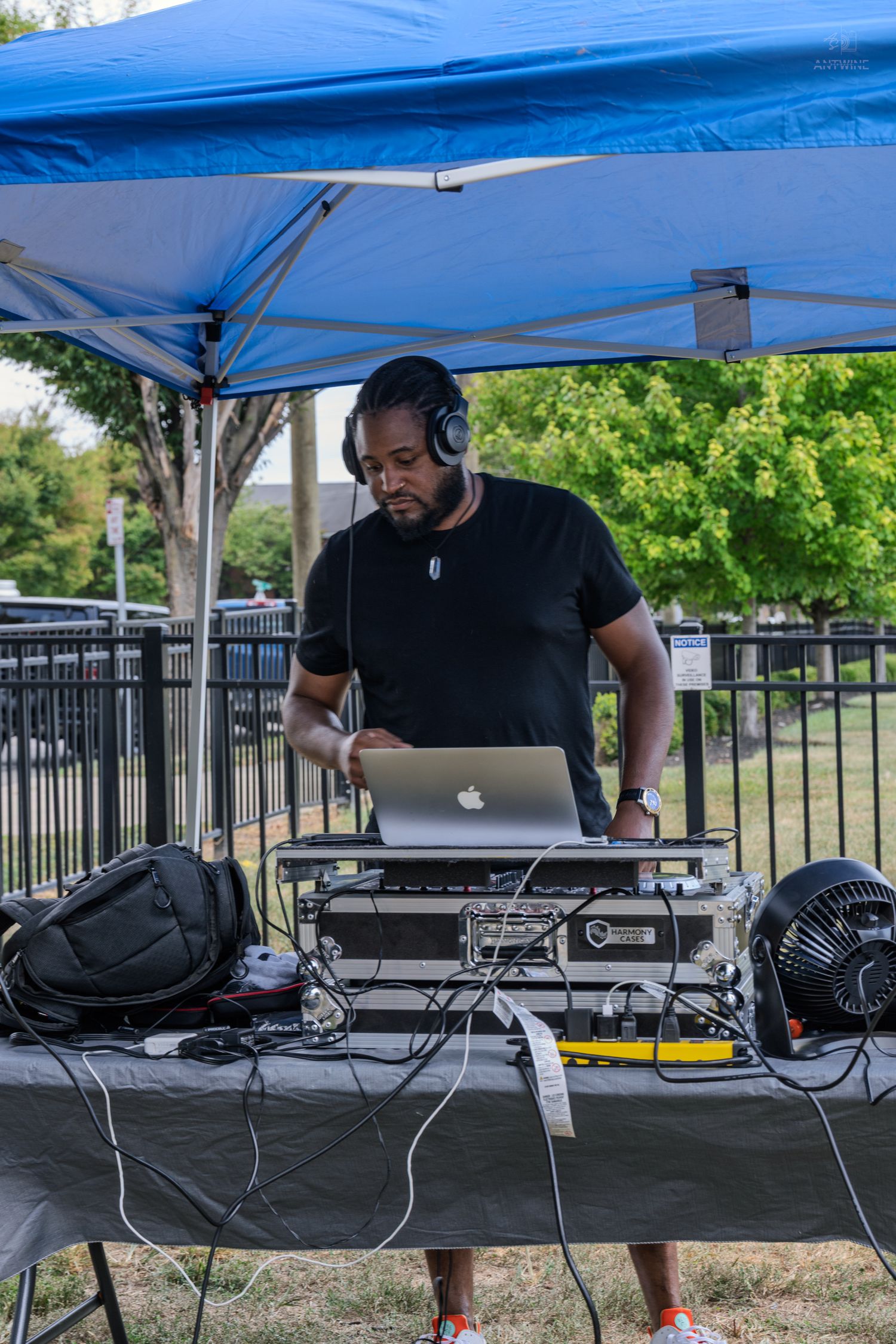 DJ in black shirt mixes music on laptop under blue canopy outdoors.