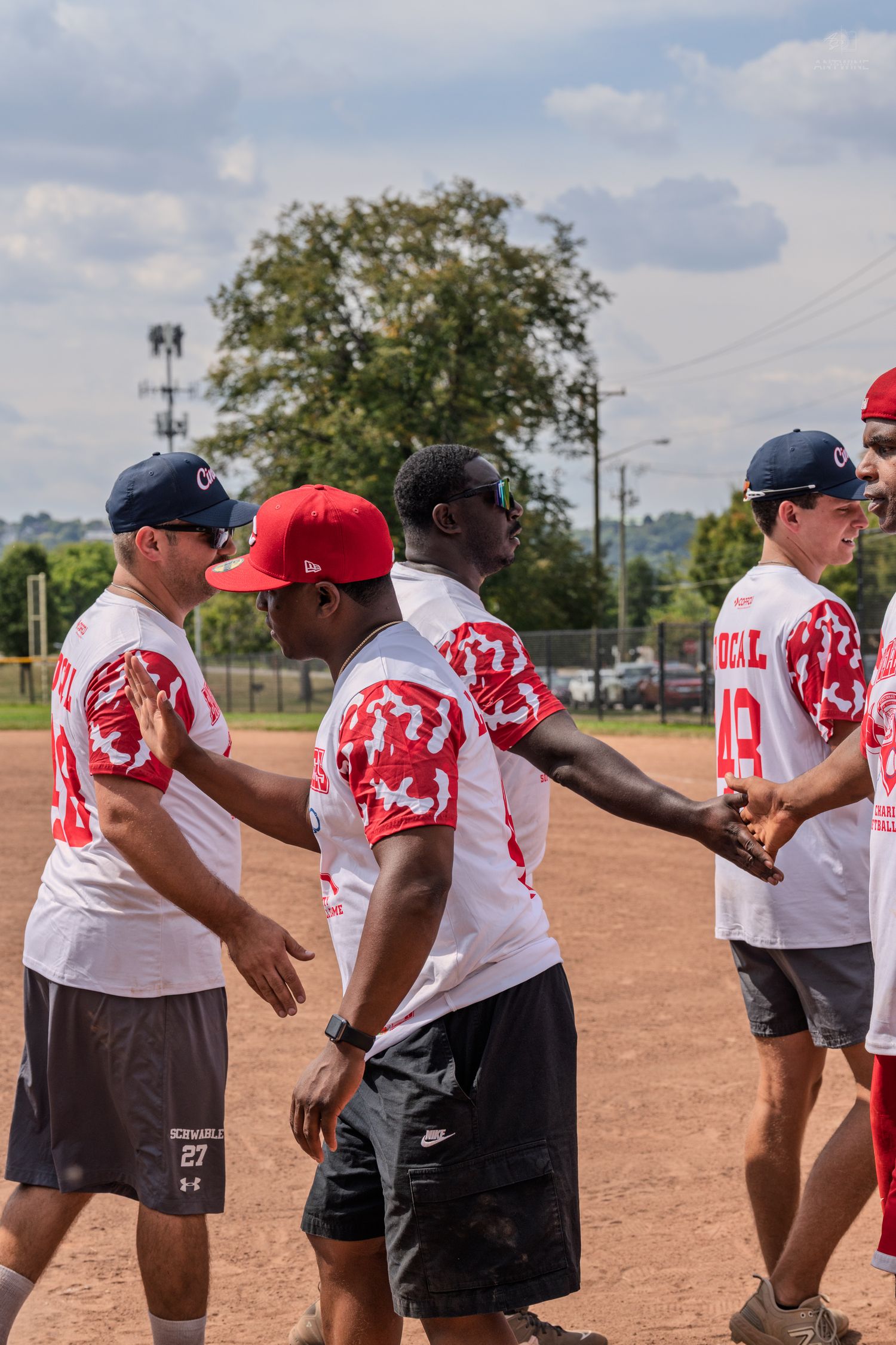Diverse softball team players shake hands on a dirt field after a game on a cloudy day.
