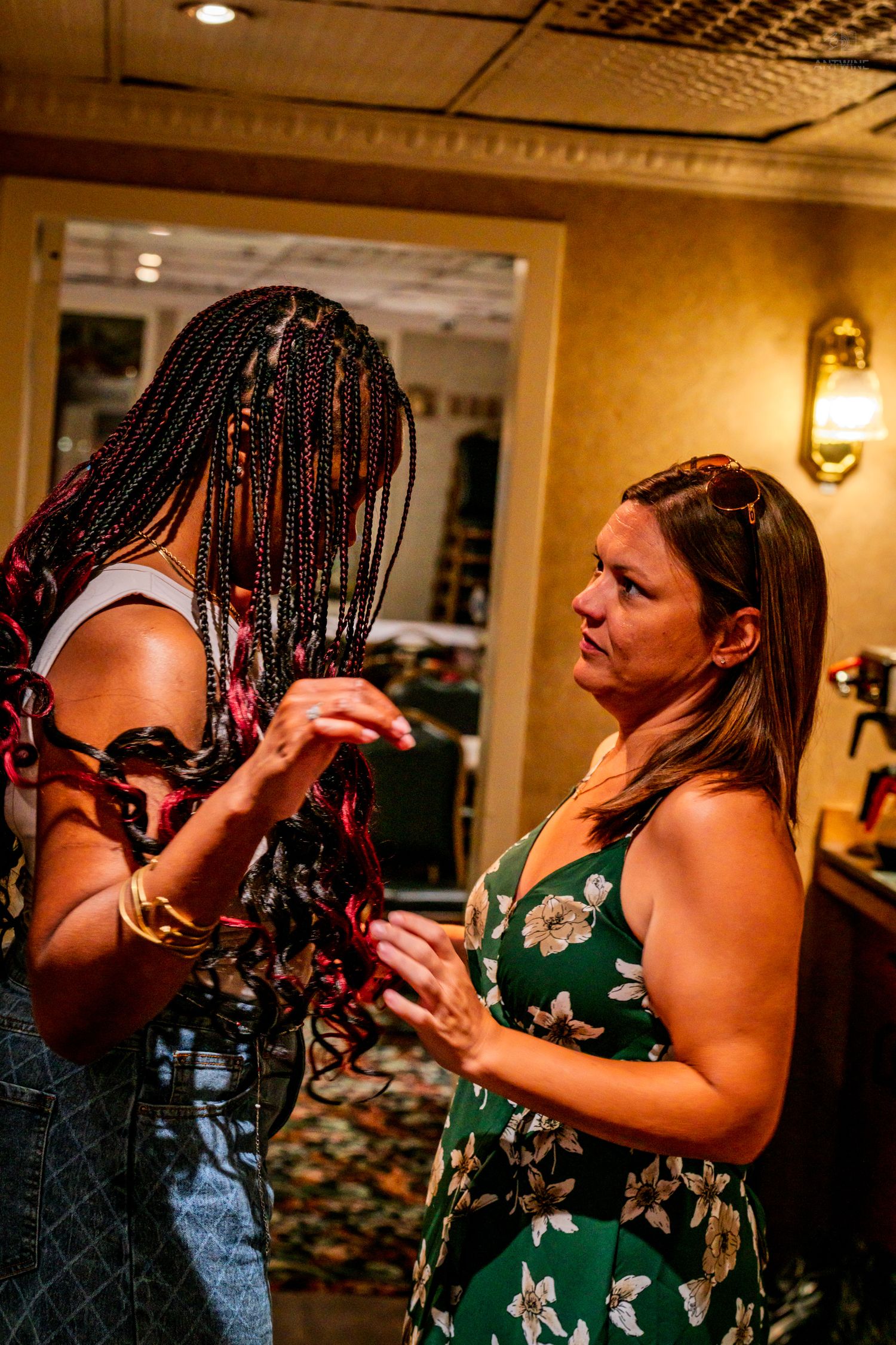 Two women converse indoors, one with long box braids and gold bracelets, the other in a green floral dress.