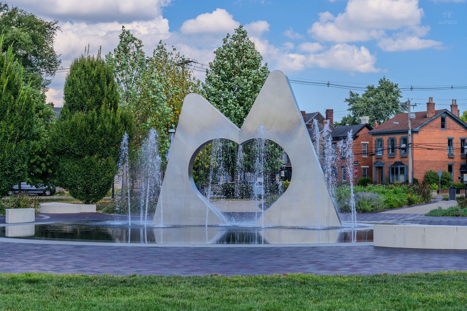 Heart-shaped fountain with water jets in a sunny urban park surrounded by green trees and brick buildings.