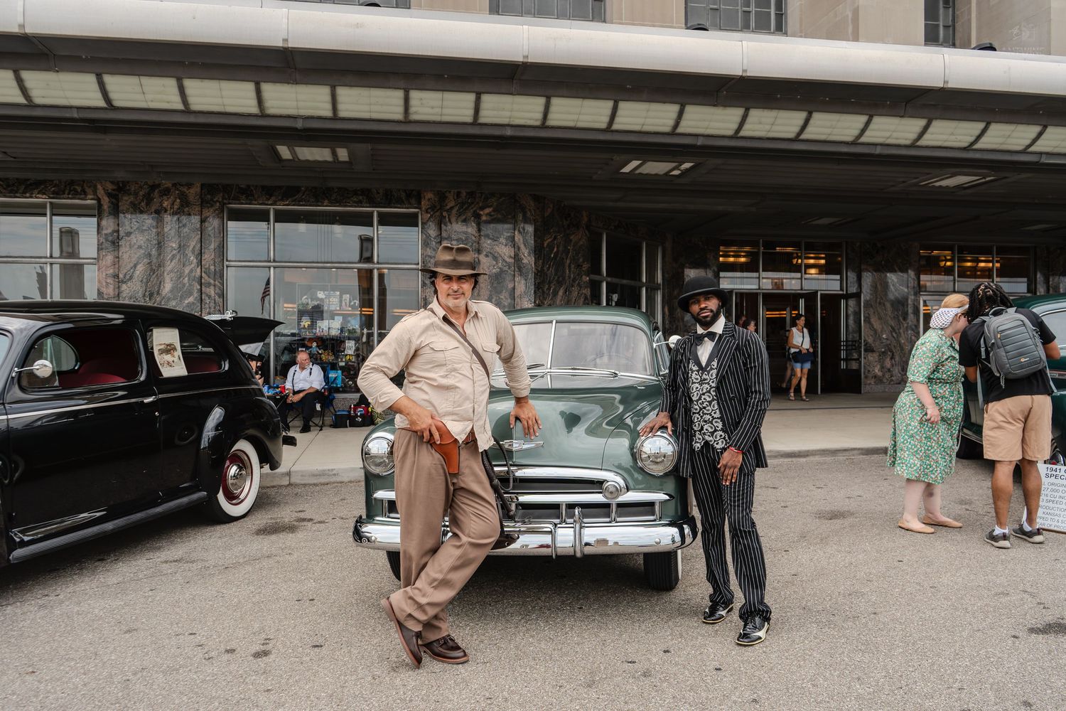 Two men in vintage attire pose beside a classic green car at a retro car show outside a historic building.