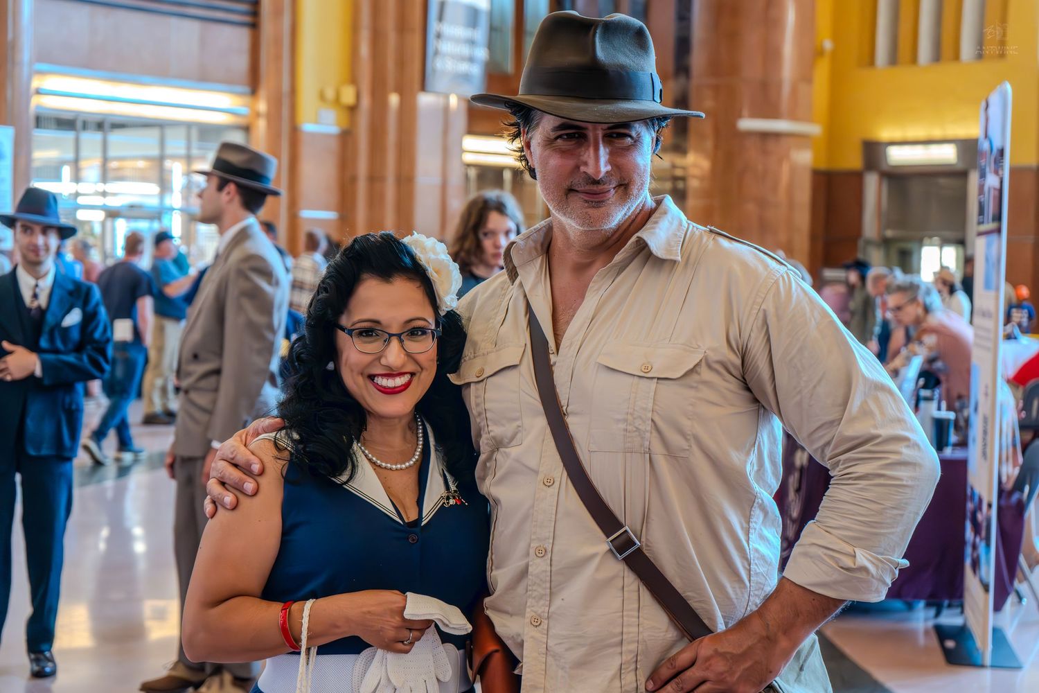 A man in Indiana Jones costume and a woman in vintage dress pose together at a retro-themed event.