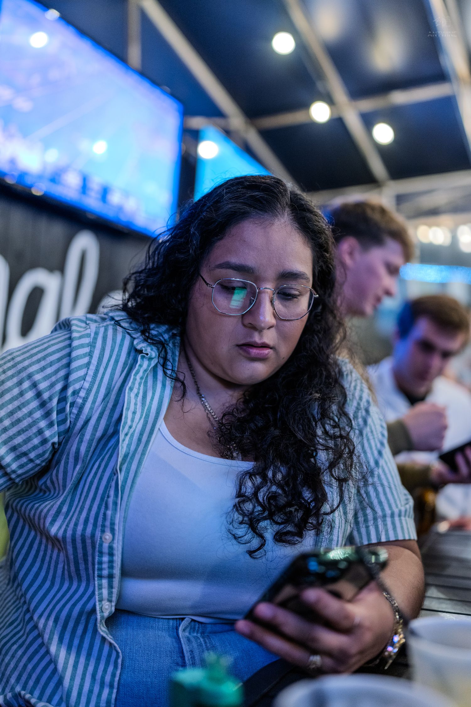Young woman with curly hair and glasses looking at her smartphone in a dimly lit bar or restaurant.