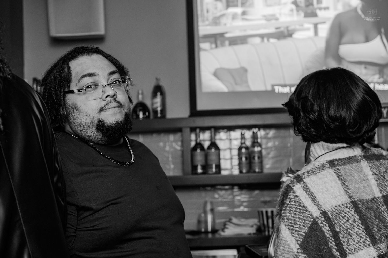 A man with glasses and a necklace sits in a bar conversing with another person, captured in black and white.