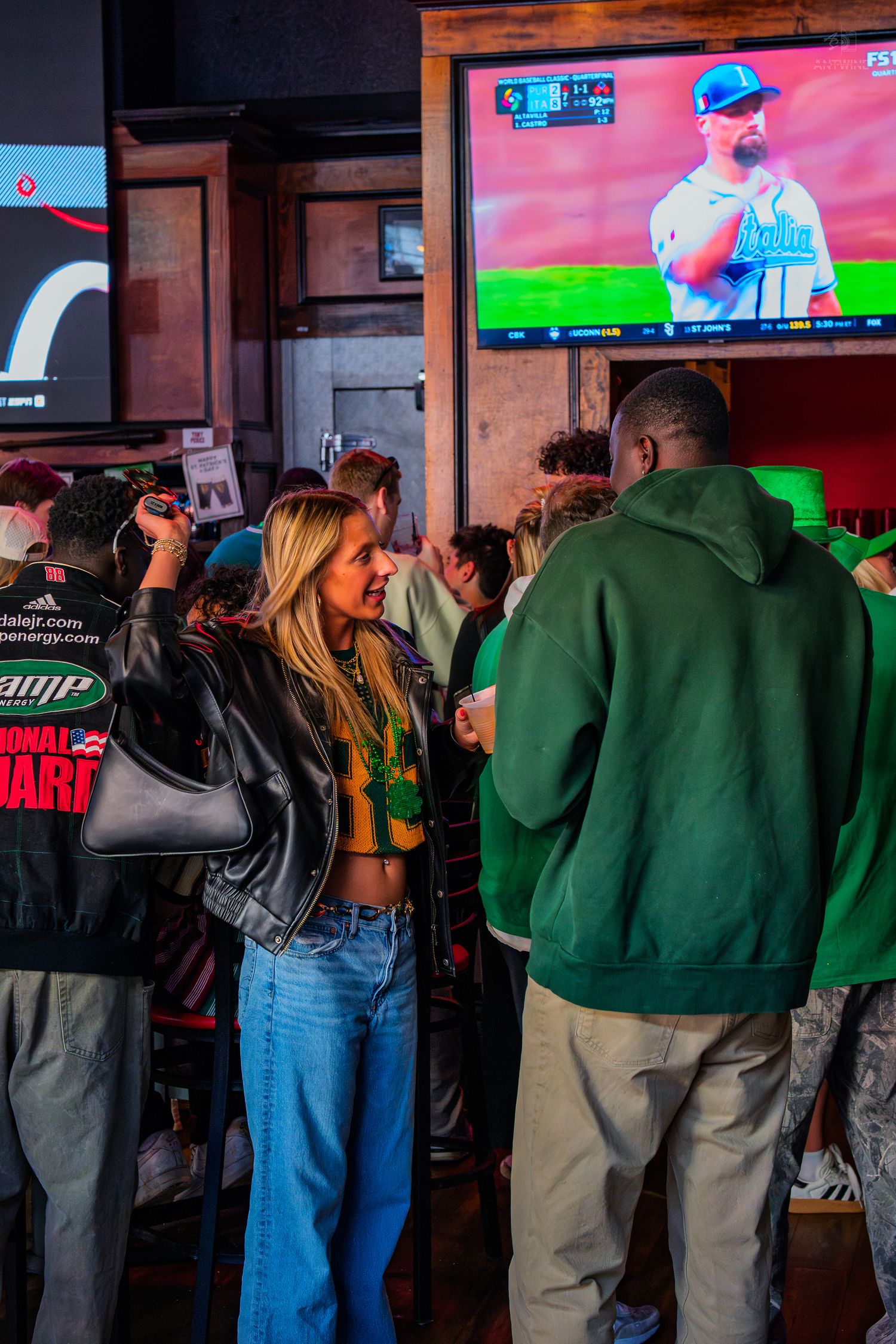 Crowd of people socializing at a sports bar with baseball game on TV screen in background.