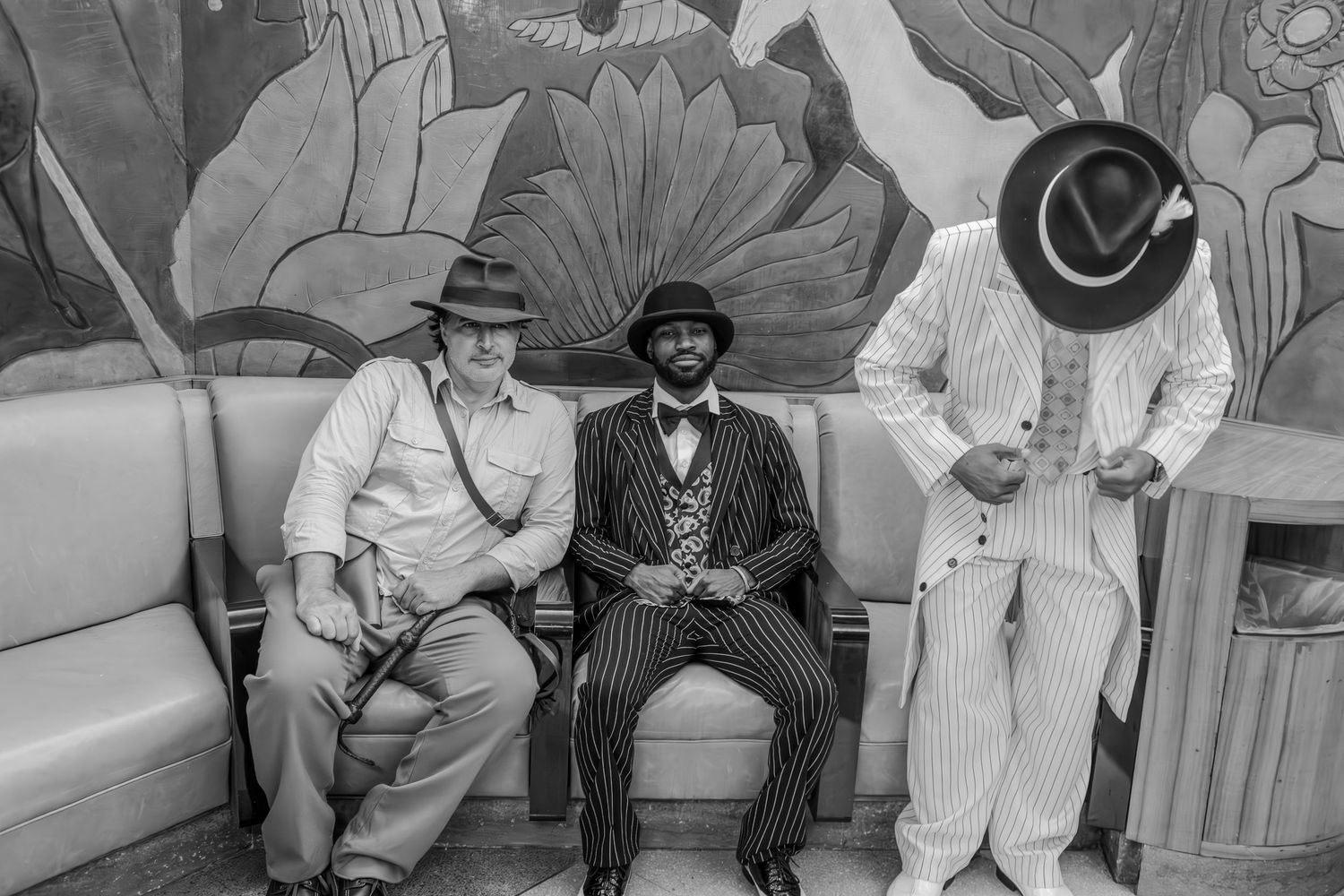 Three men in stylish vintage suits and fedora hats pose together in a black and white photo before a floral mural.