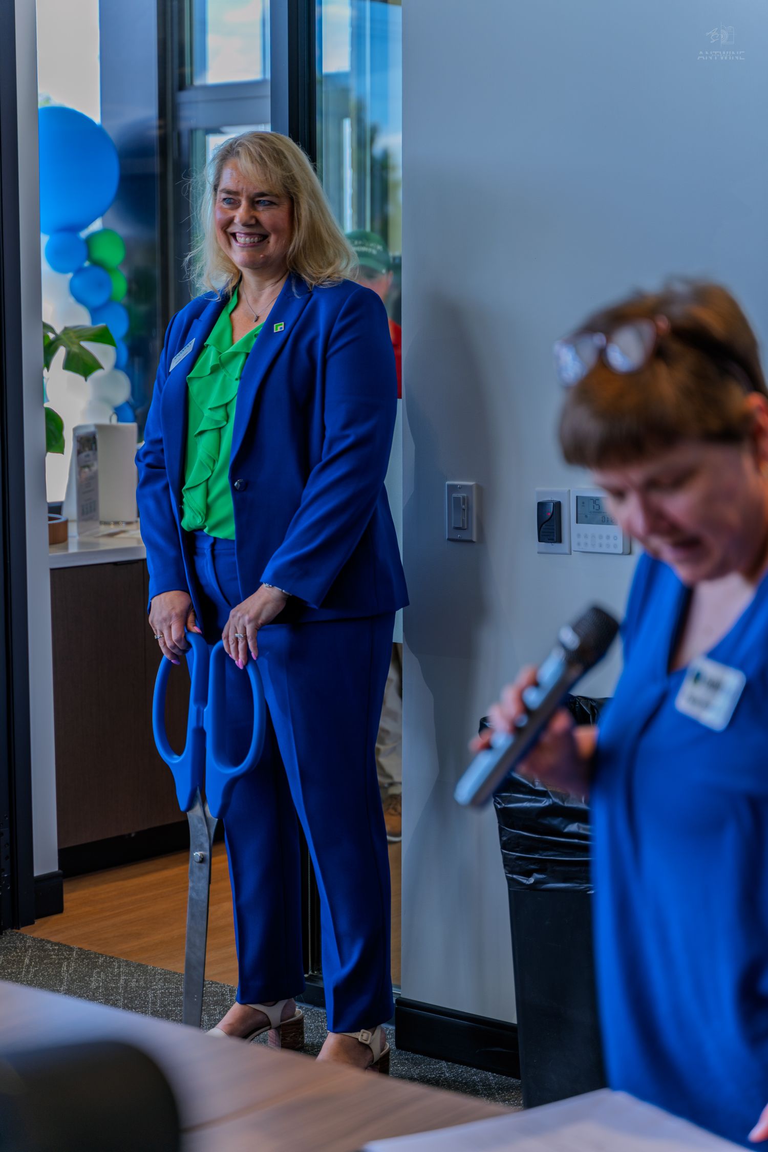 Two women in blue suits at a ribbon-cutting ceremony, one holding large scissors, with blue and green balloons in background.