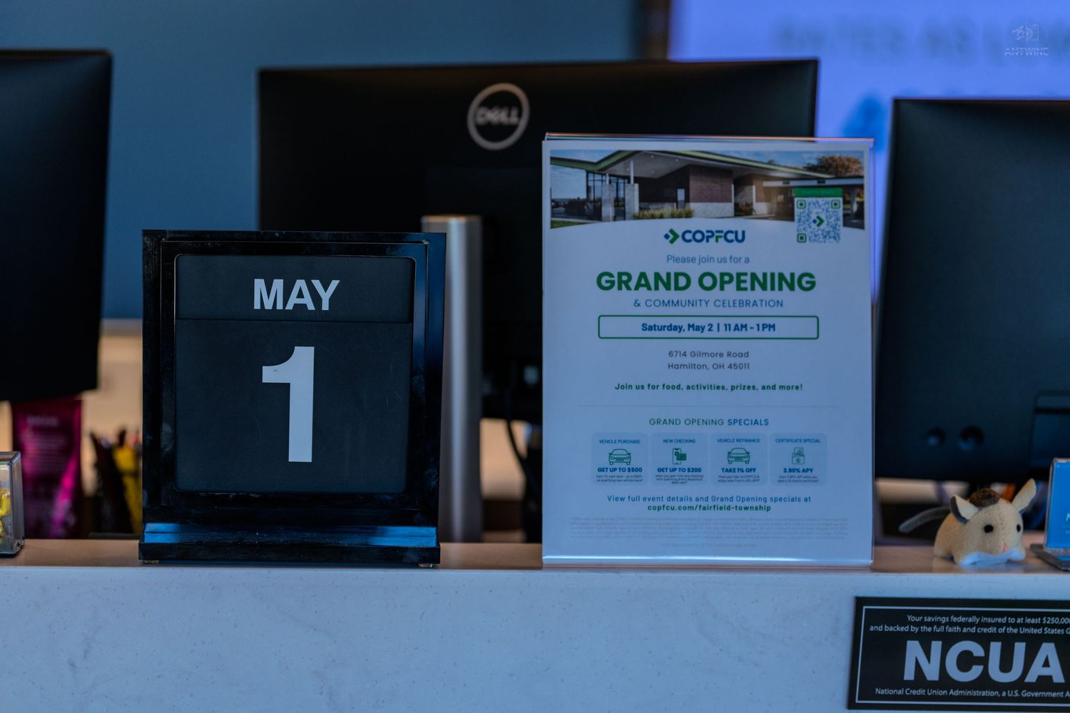 COPFCU credit union grand opening flyer on desk with May 1st calendar, Dell monitors, and NCUA sign in background.