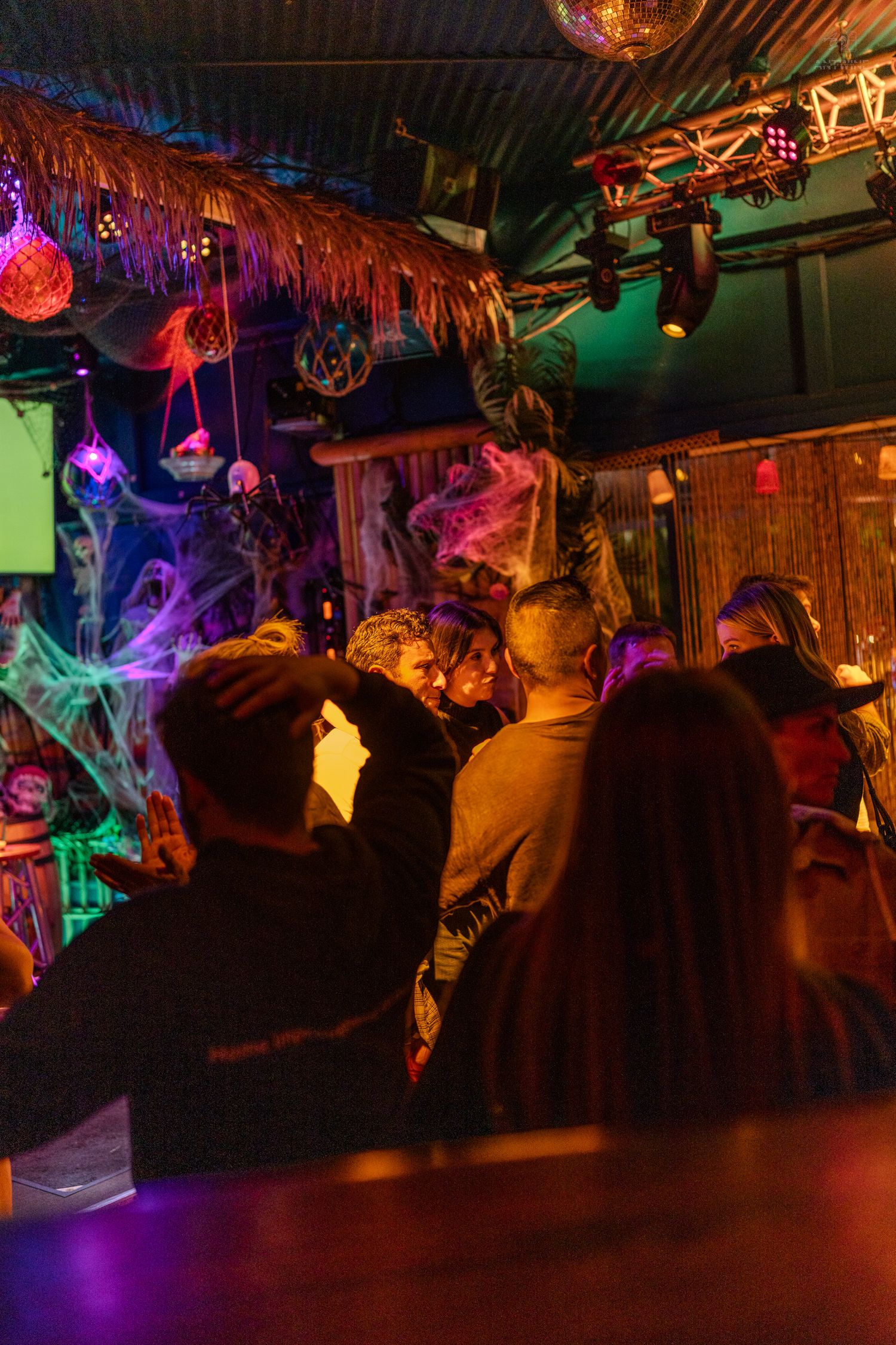 Crowd enjoying a Halloween-themed tiki bar with colorful neon lights and spooky cobweb decor.