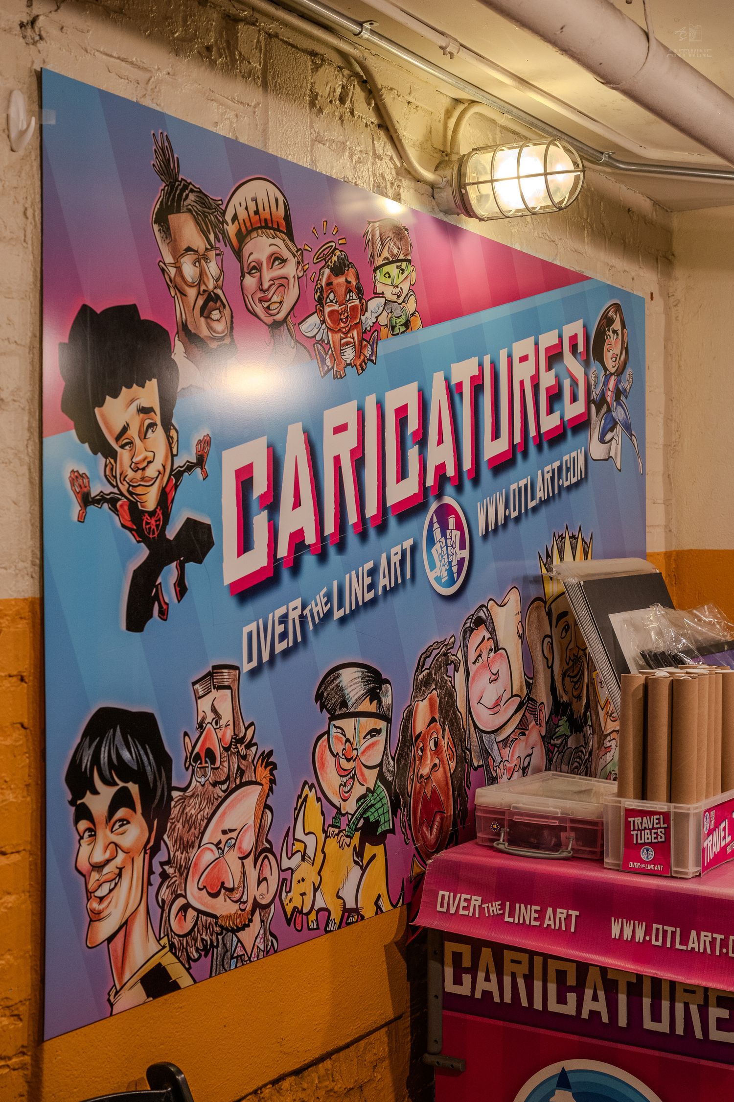 Colorful caricatures promotional sign for Over The Line Art displayed on a brick wall with travel tubes nearby.