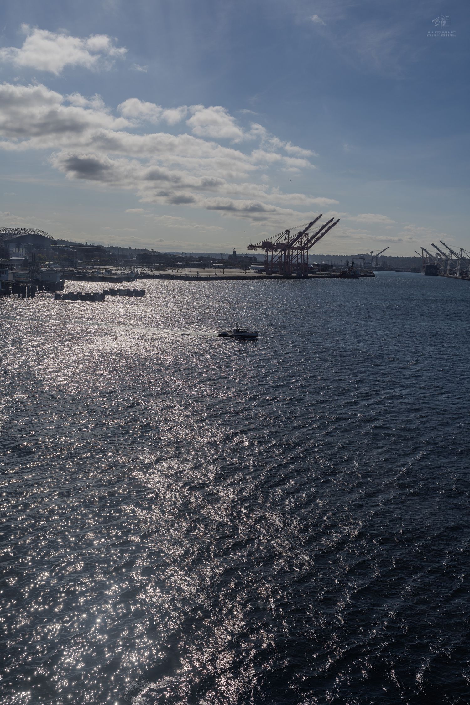 Seattle harbor with industrial port cranes in the distance and a small boat crossing the sunlit Puget Sound waters.