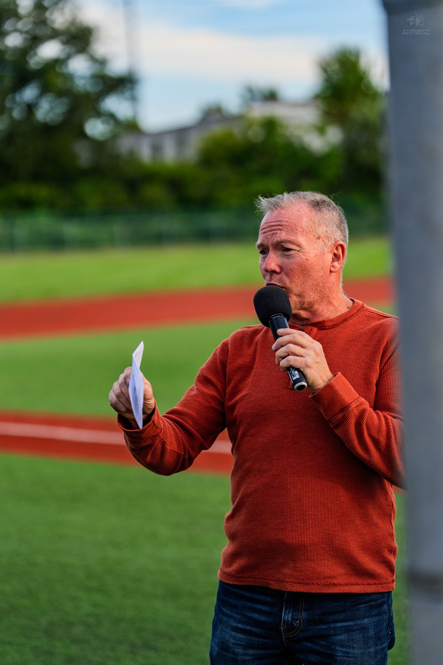Older man in red sweater speaks into microphone while reading from paper at outdoor sports event.