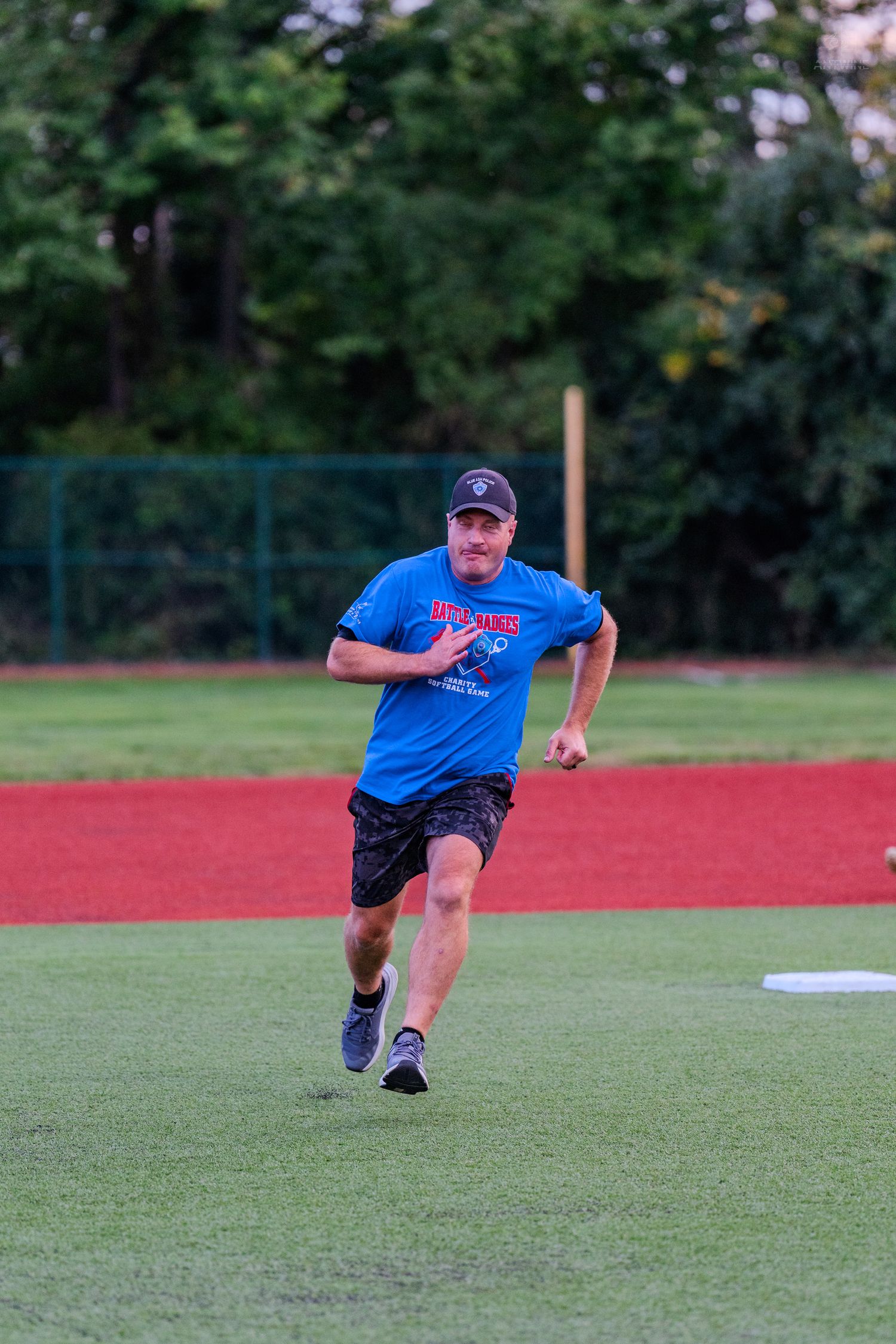 Man in blue shirt and cap running on a baseball field with green grass and red dirt track.