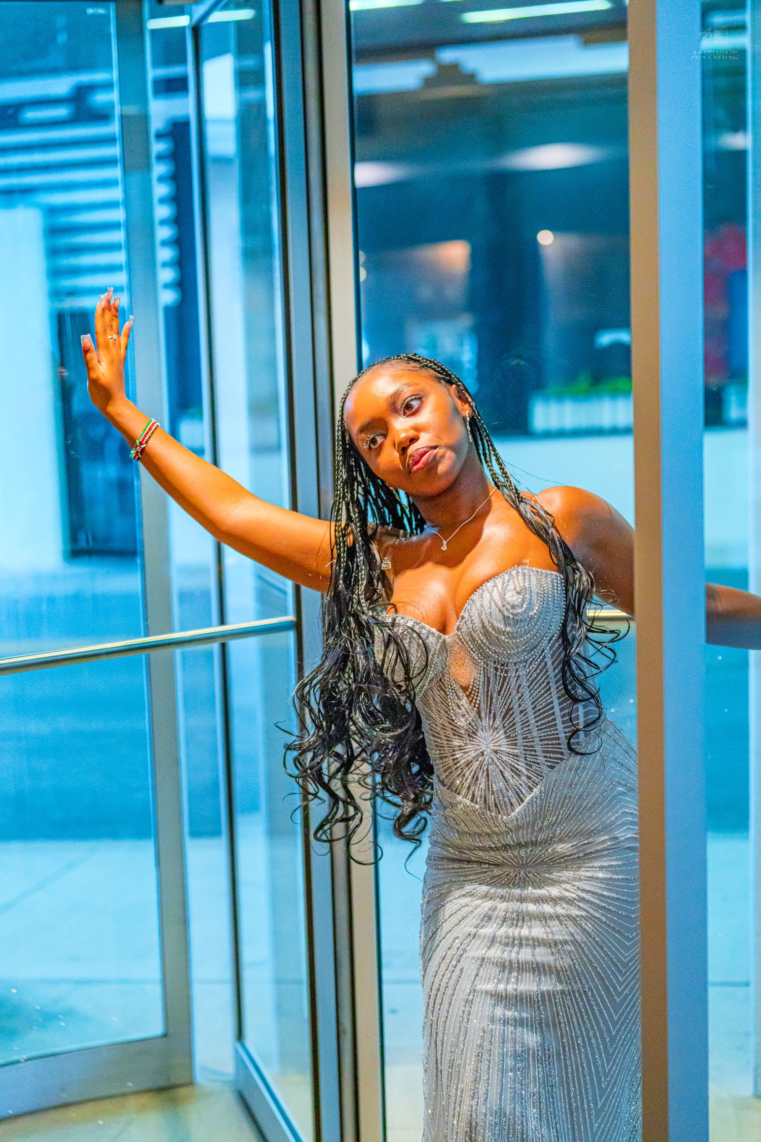Young woman in silver beaded gown posing by glass doors with long braided hair.