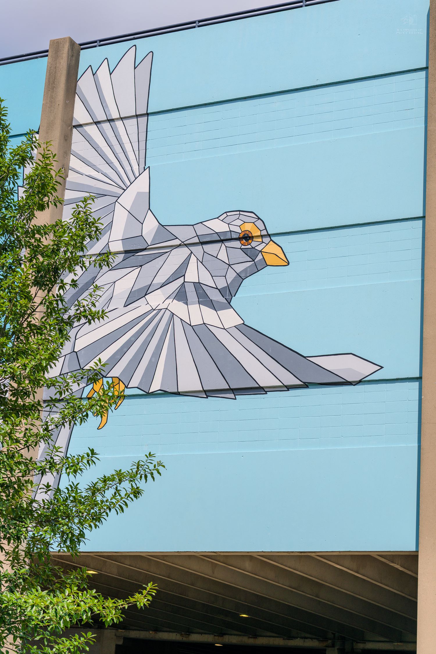 Geometric low-poly style white bird mural painted on the exterior wall of a modern building against a blue sky.