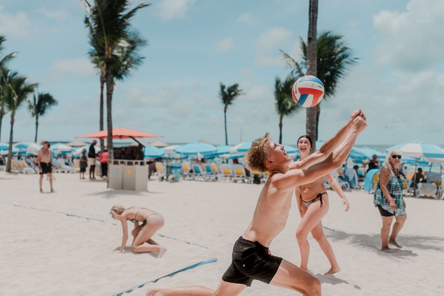 Shirtless players jumping to spike a volleyball on a crowded tropical beach.