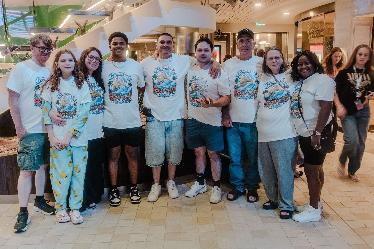 Group of nine cruise passengers posing together in matching white tropical graphic t-shirts indoors.