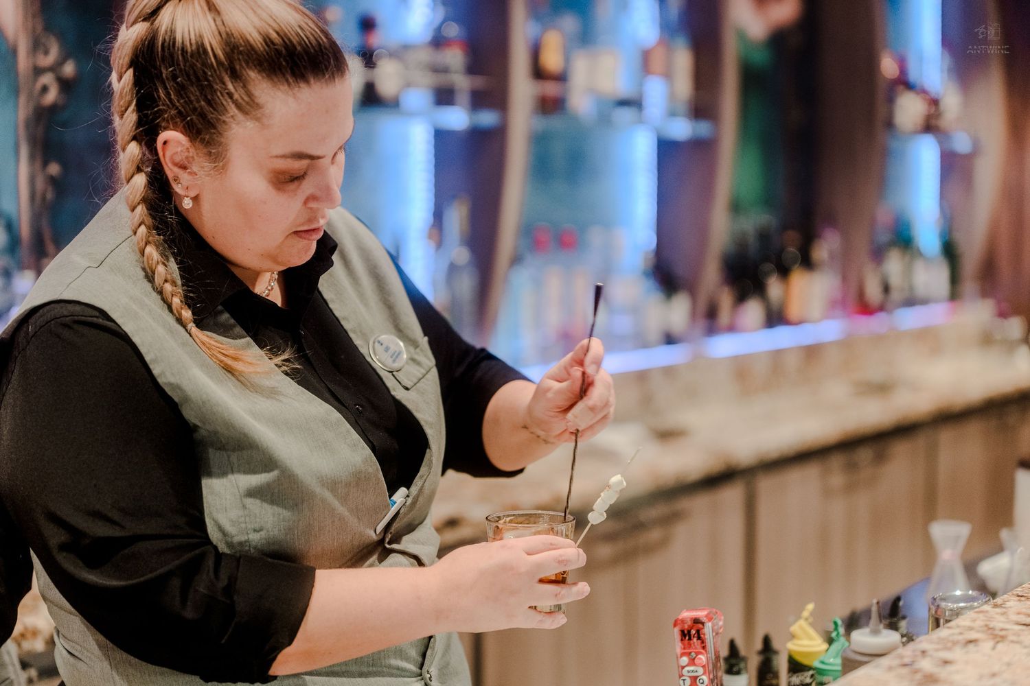 Female bartender in apron carefully mixing a cocktail with a spoon at a dimly lit bar.