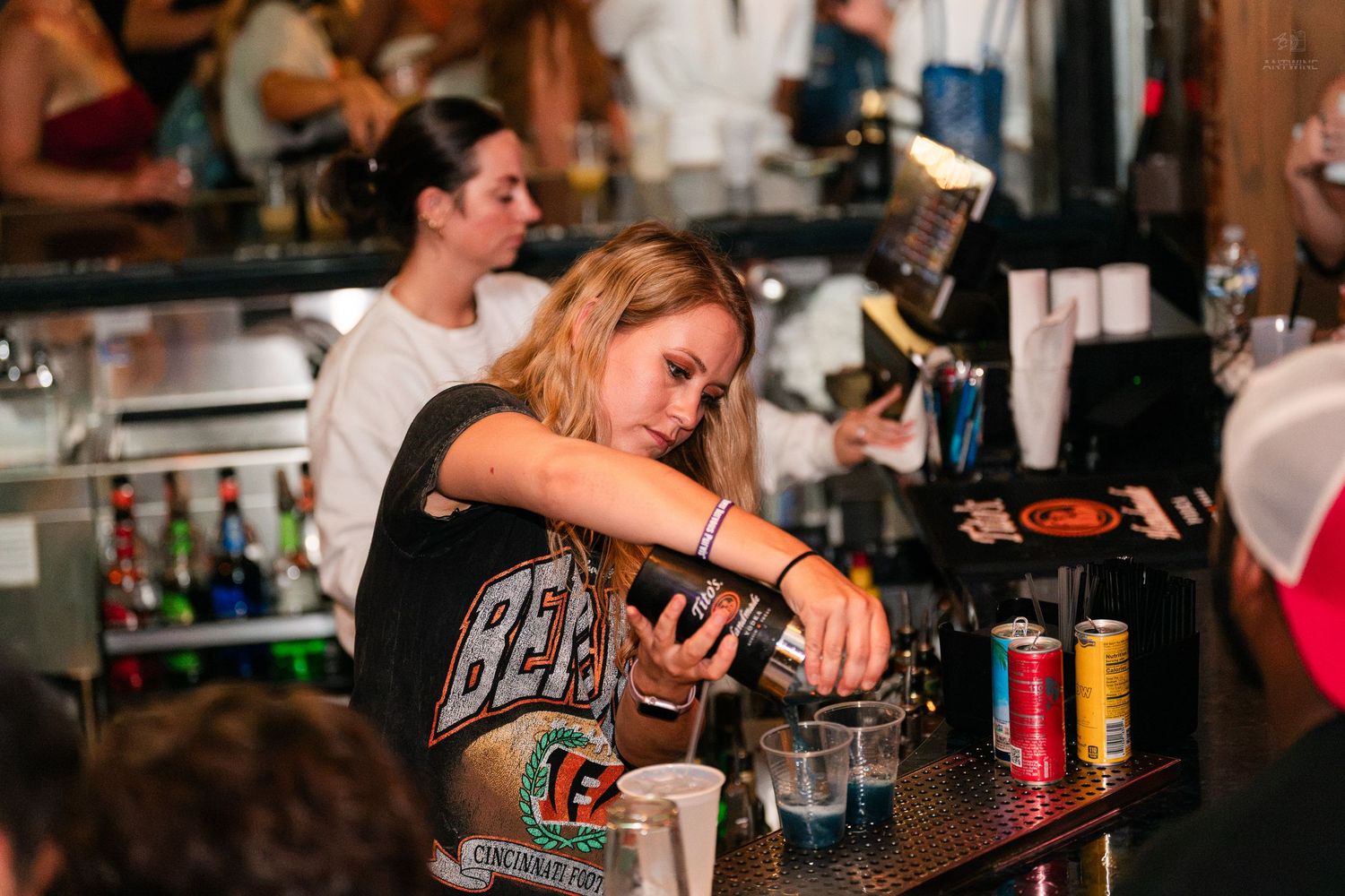 Blonde female bartender pouring drinks at a crowded bar wearing a black graphic tee.