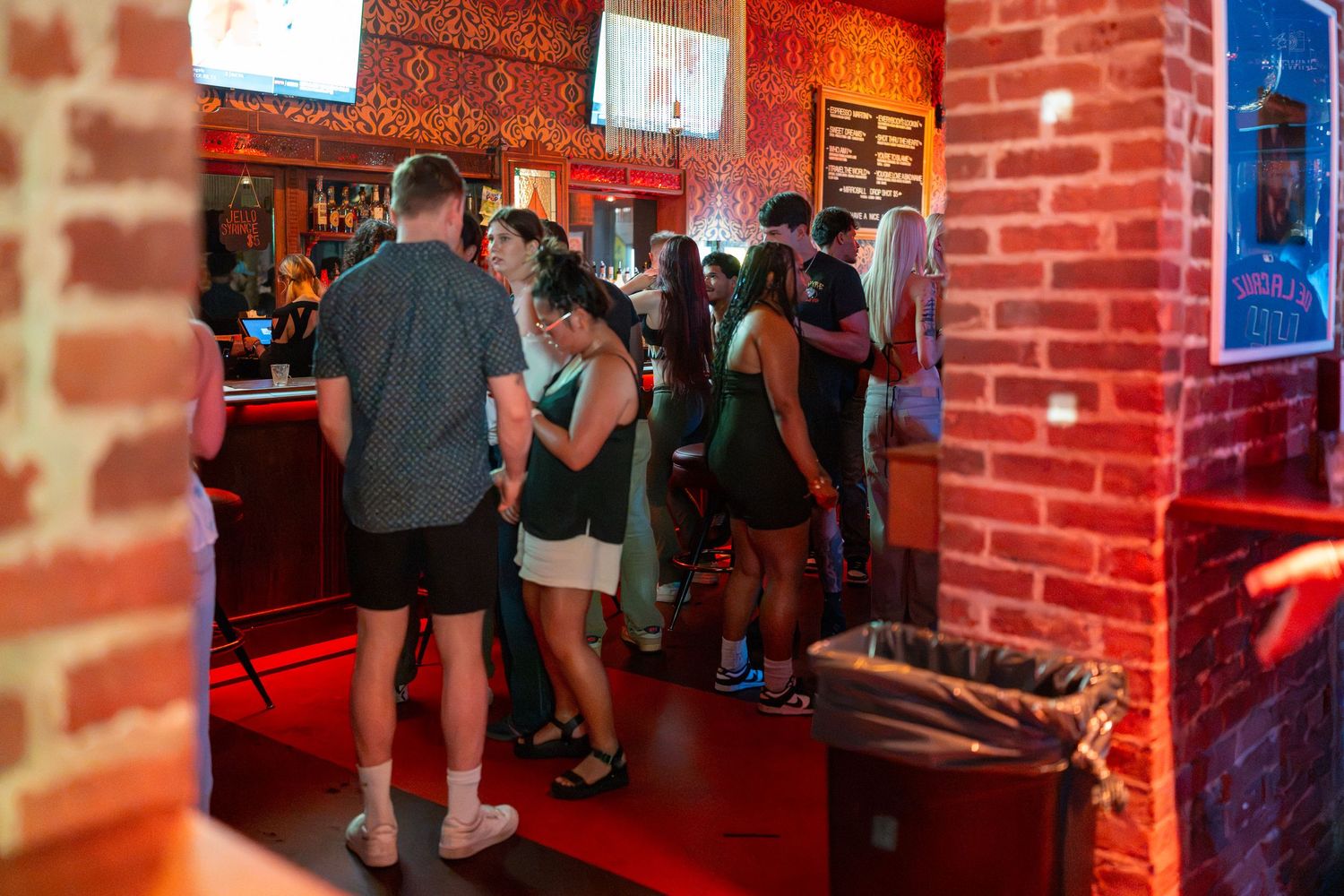 Crowded bar interior with brick walls, red lighting, and groups of people socializing near the counter.