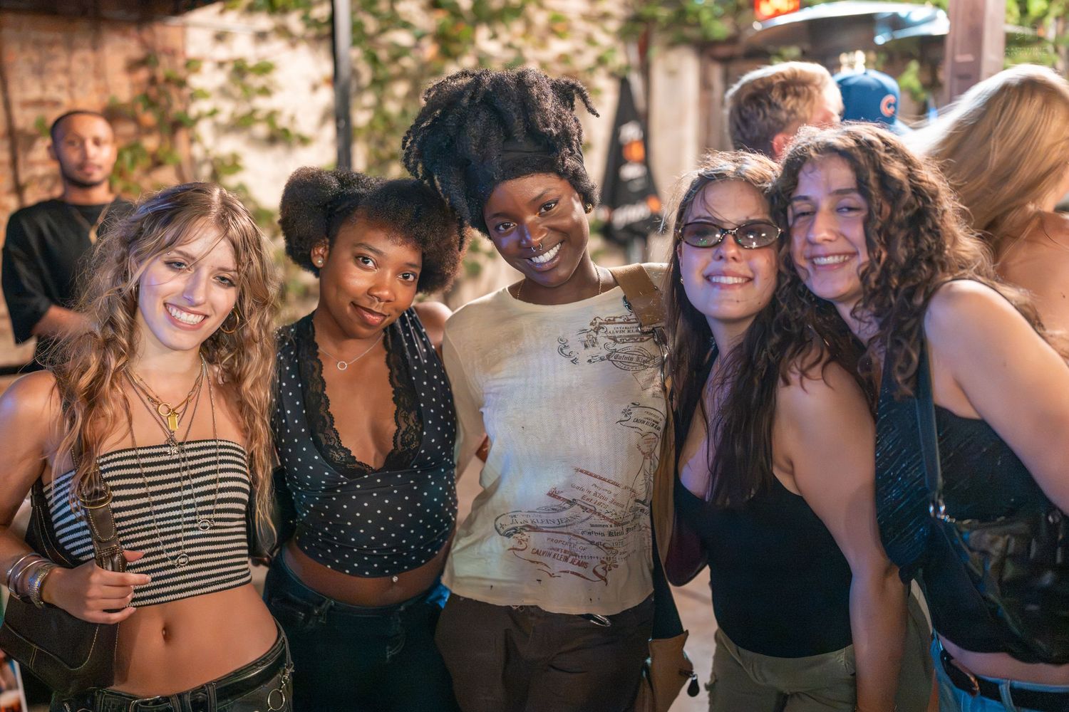 Five diverse young women smile and pose together at an outdoor social gathering at night.