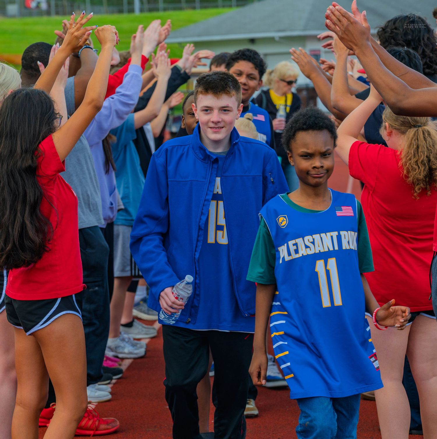 Two young athletes in blue Pleasant Bay jerseys walk through a high-five tunnel of cheering peers on a track.