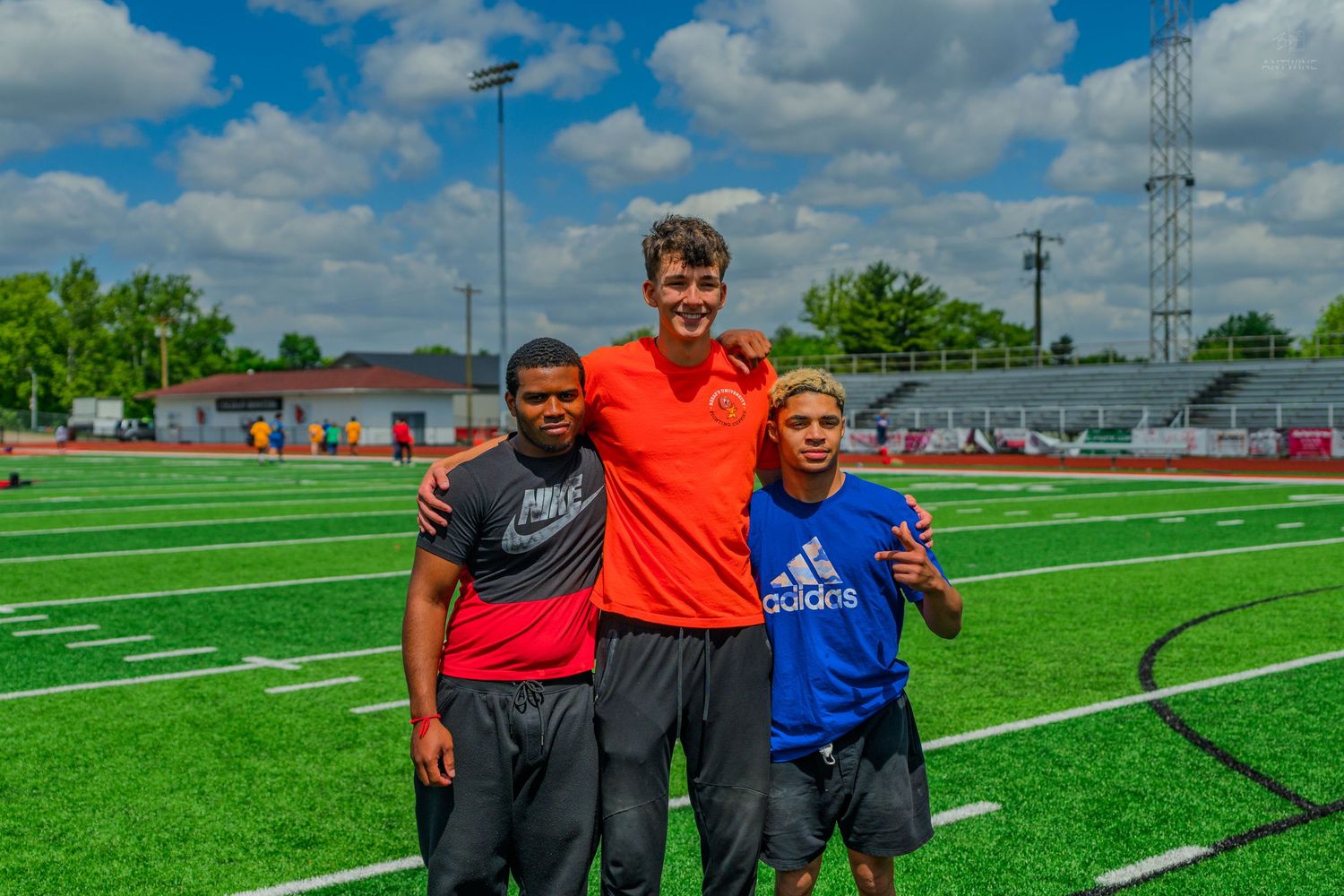 Three young football players posing together on a green turf field during an outdoor sports camp on a sunny day.