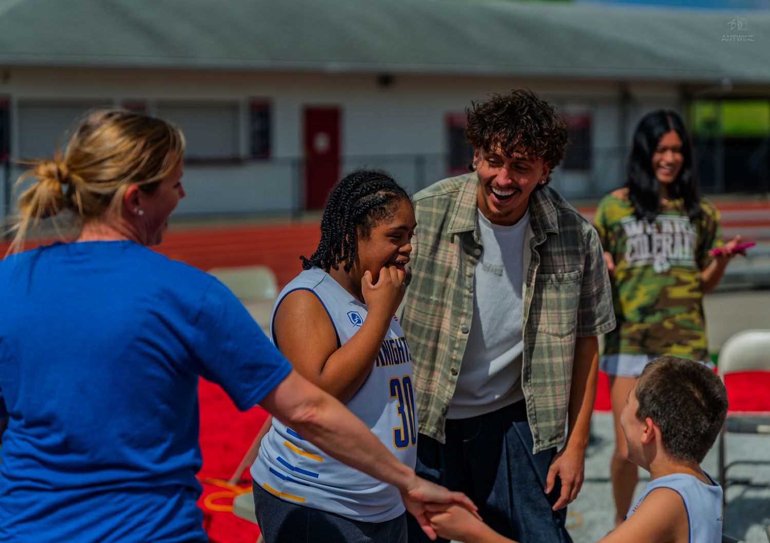 Group of people smiling and interacting outdoors at a track and field event on a sunny day.