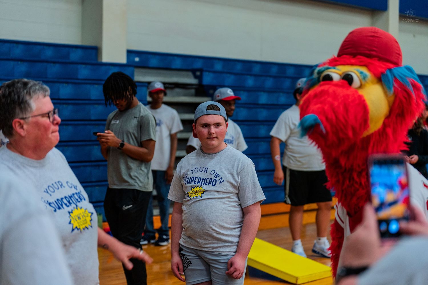 Young man poses with a red mascot costume character in a school gymnasium during a special event.