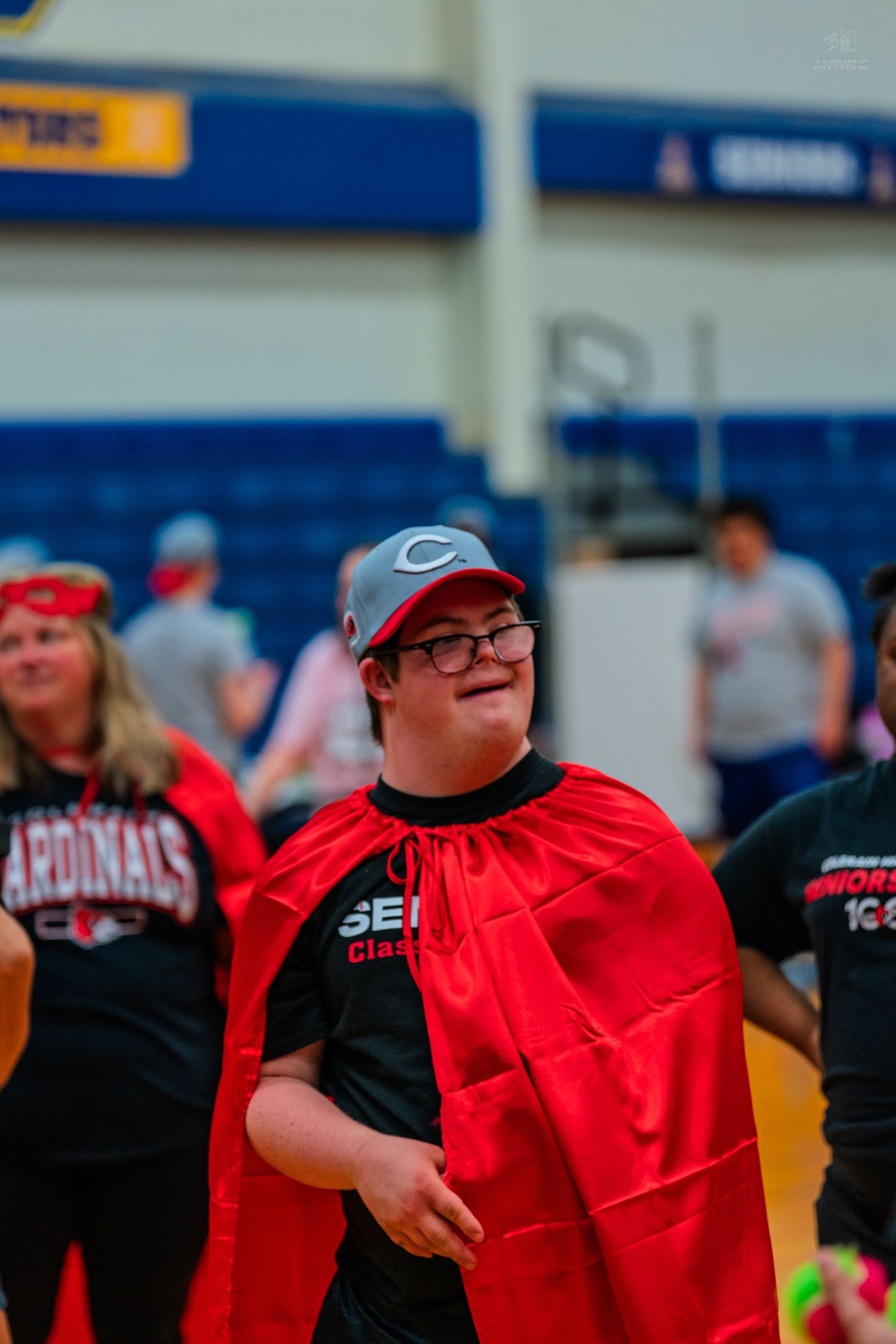 Young man with Down syndrome wearing red cape and Cardinals shirt smiles at indoor sports event.
