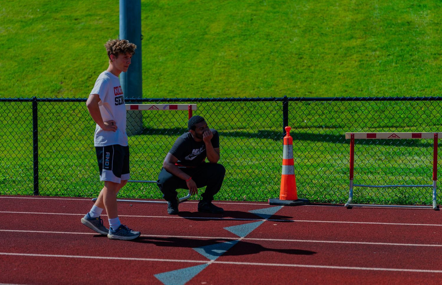 Coach crouching on track guiding young athlete at starting line near orange cone and green field.