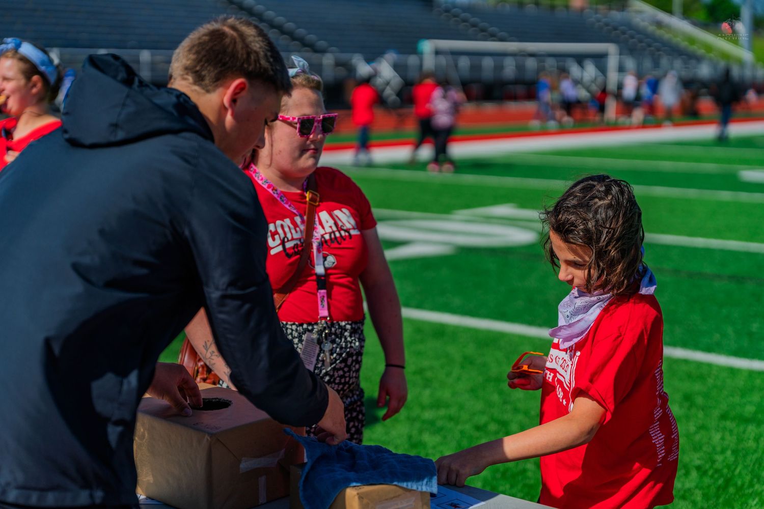 Adults and child interact at an outdoor football field event, wearing red shirts on a sunny day.