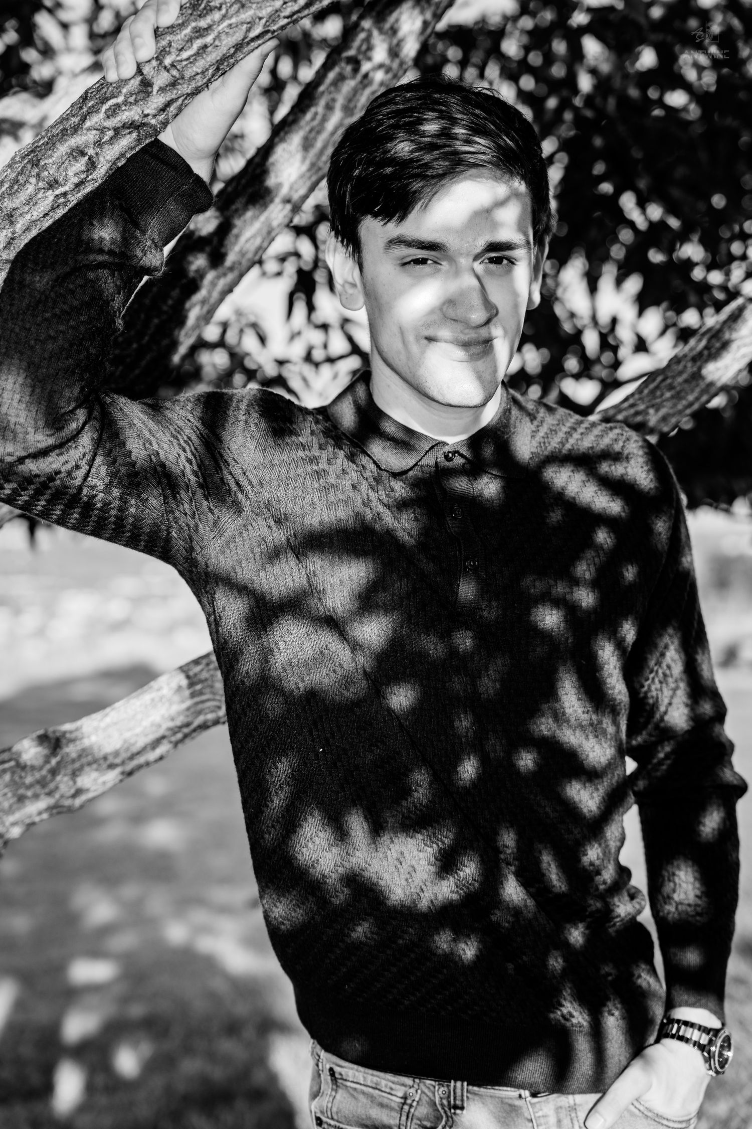 Black and white portrait of a smiling young man leaning against a tree with dappled sunlight shadows on his sweater.