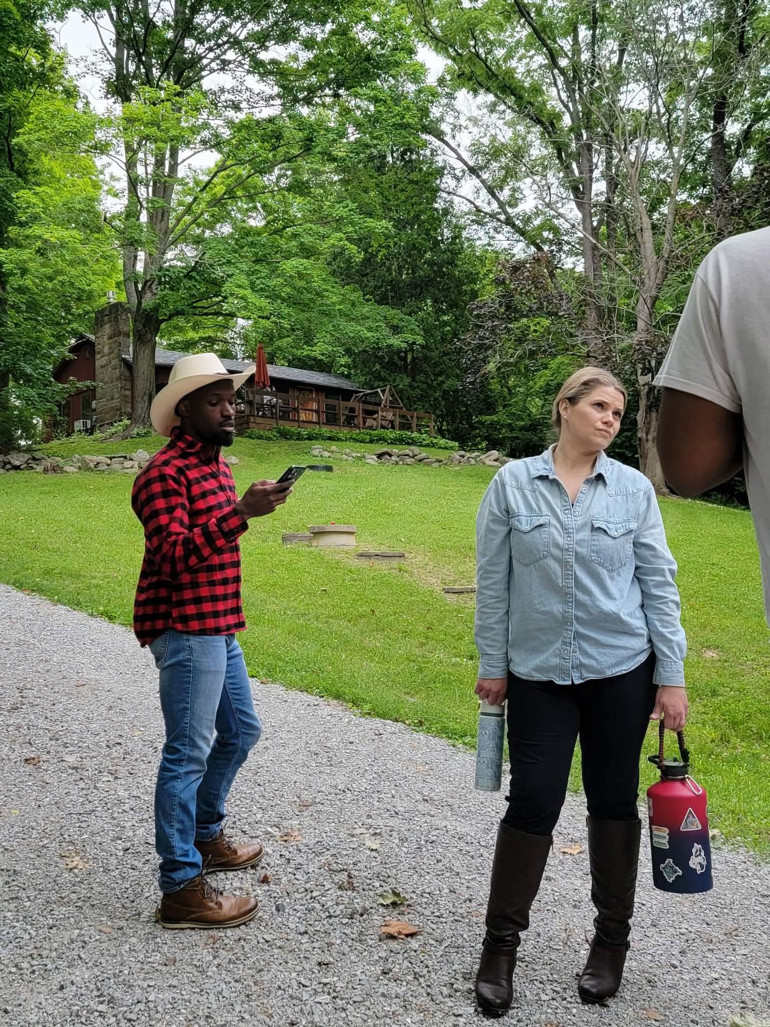 Woman in denim shirt holding water bottle stands on gravel path near man in red plaid shirt and cowboy hat outdoors.