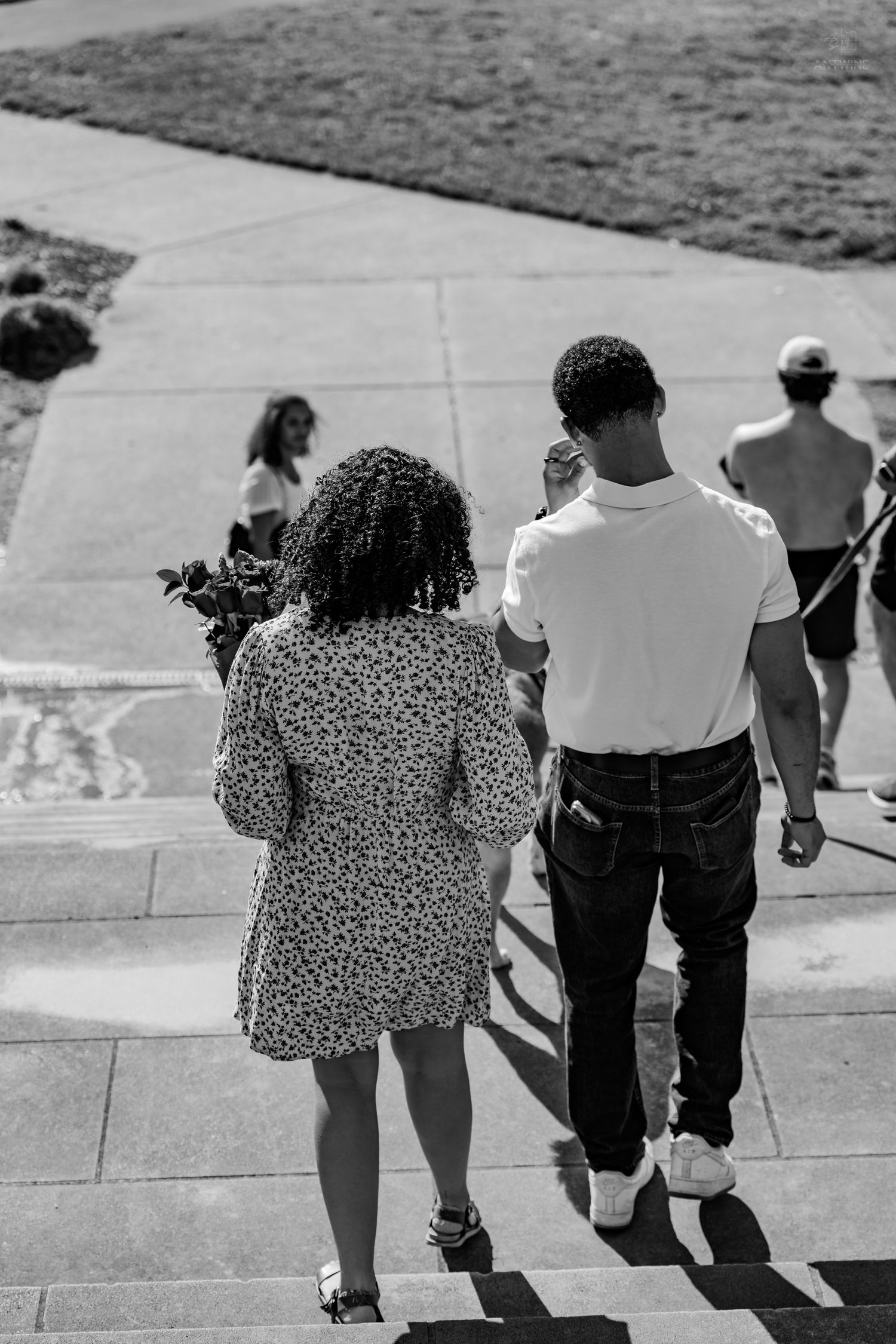 Aerial black and white view of a couple walking down steps, woman holding flowers in a floral dress.