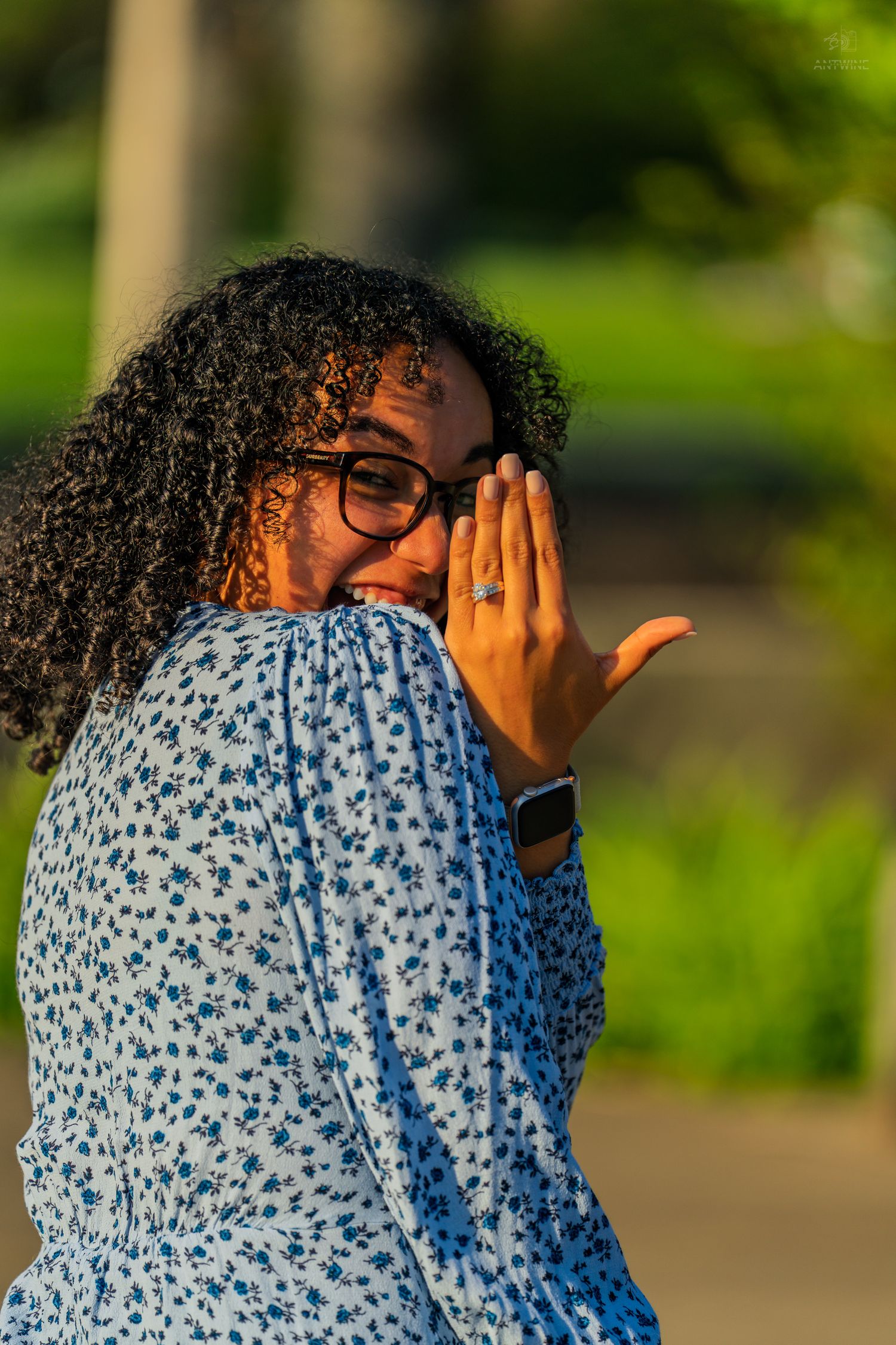Joyful woman in floral dress with curly hair and glasses shows off engagement ring outdoors.