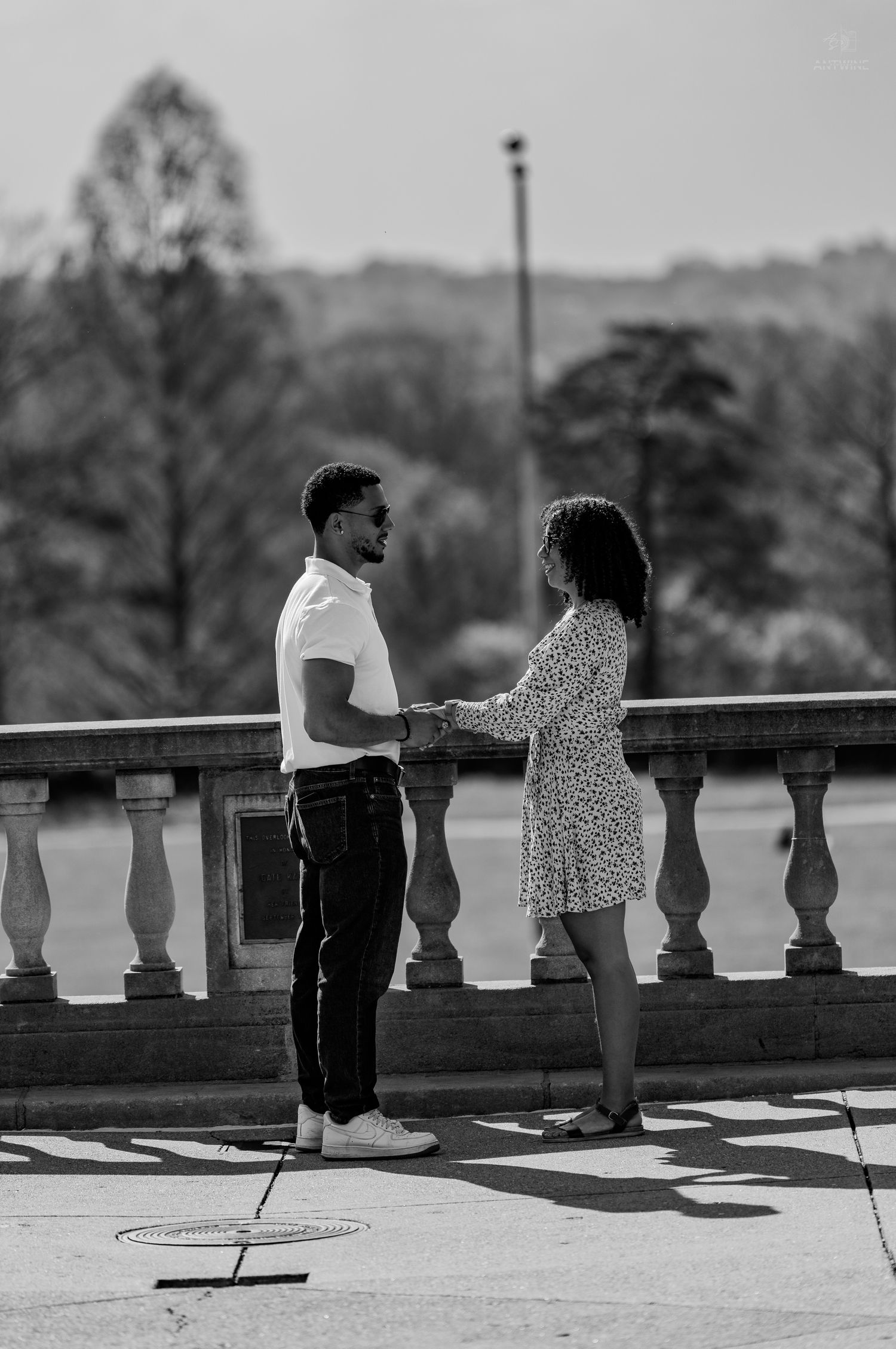 Black and white photo of a couple holding hands on a sunny terrace with park backdrop.