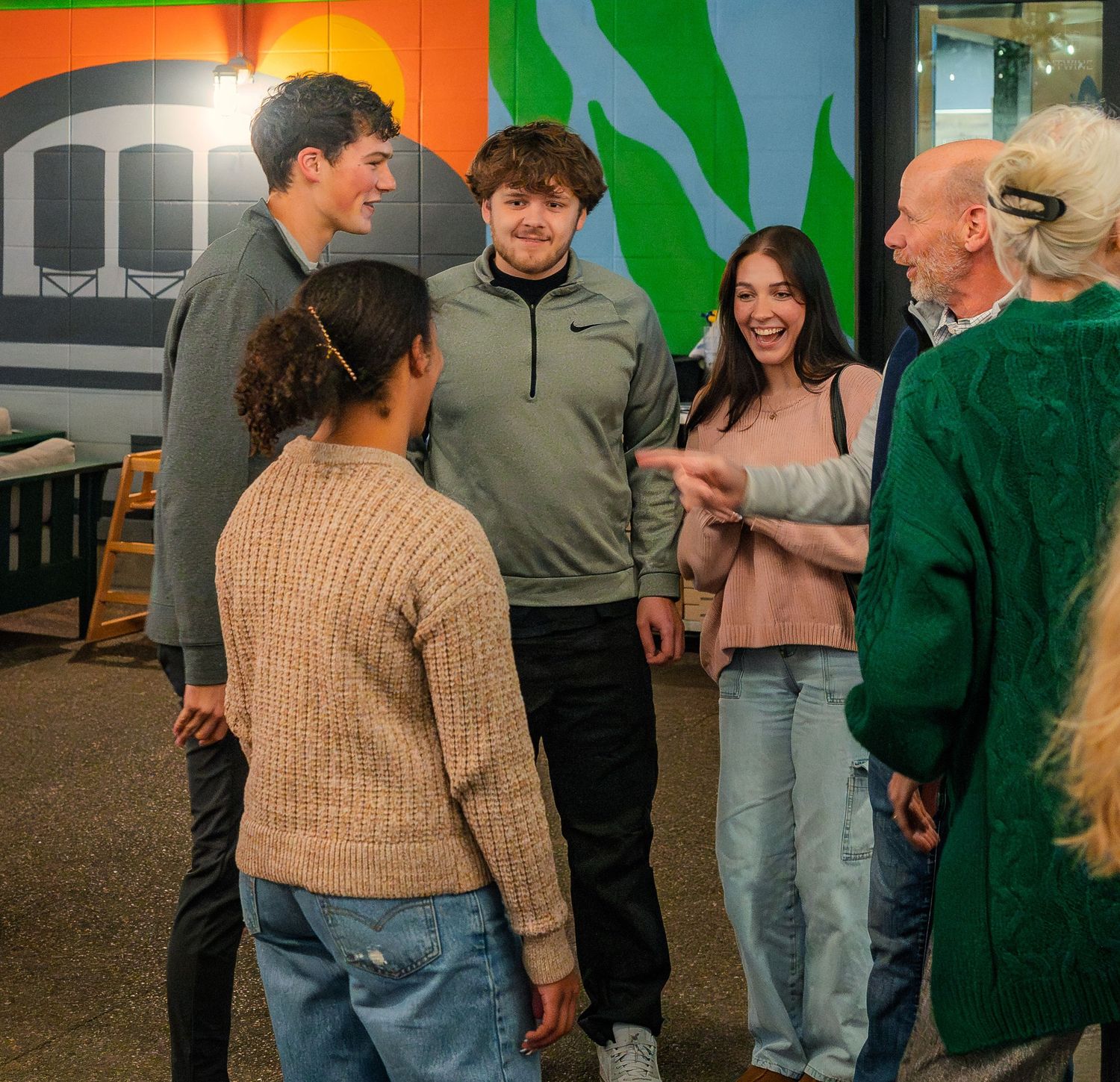 Group of young adults laughing and conversing with an older woman inside a brewery.