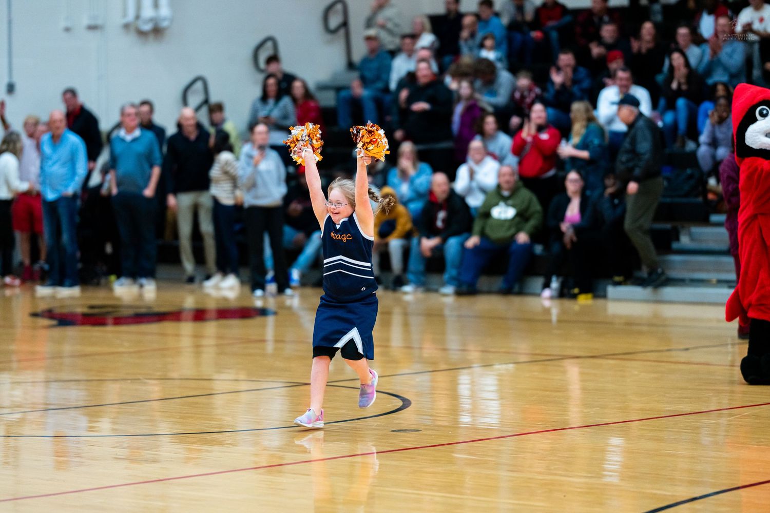 Young cheerleader in navy uniform raises gold pom-poms on basketball court with mascot visible nearby.