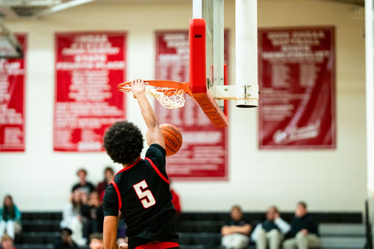 Player #5 completes powerful slam dunk through the net during high school basketball game.