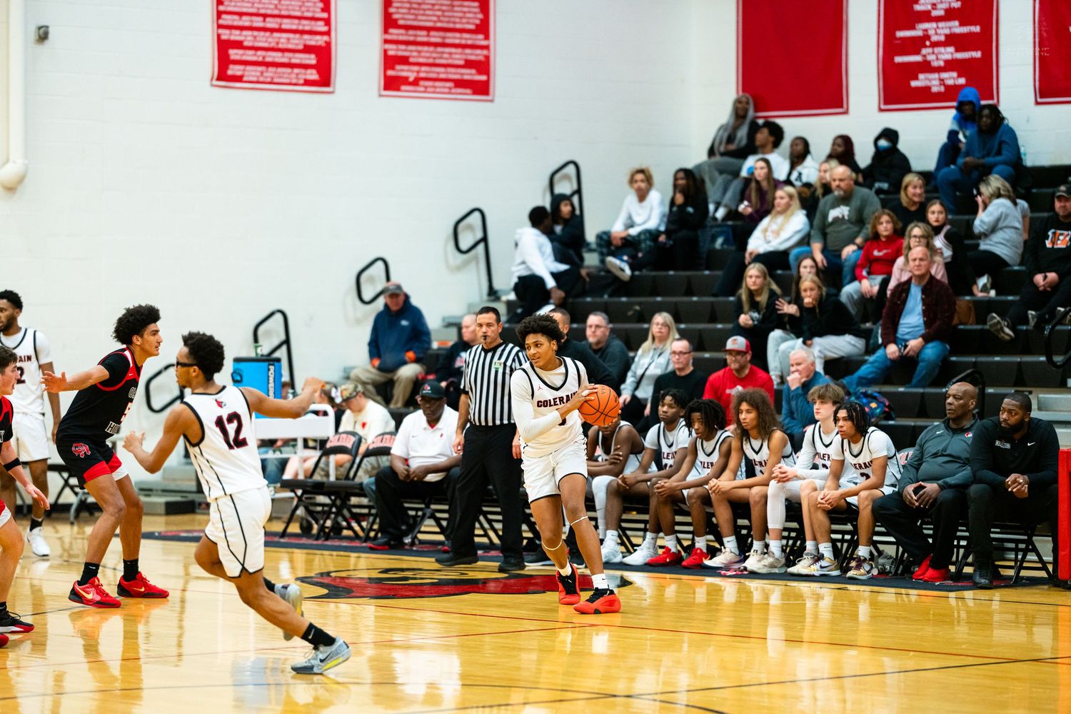 High school basketball players in white uniforms move down the court during an indoor game with spectators watching.