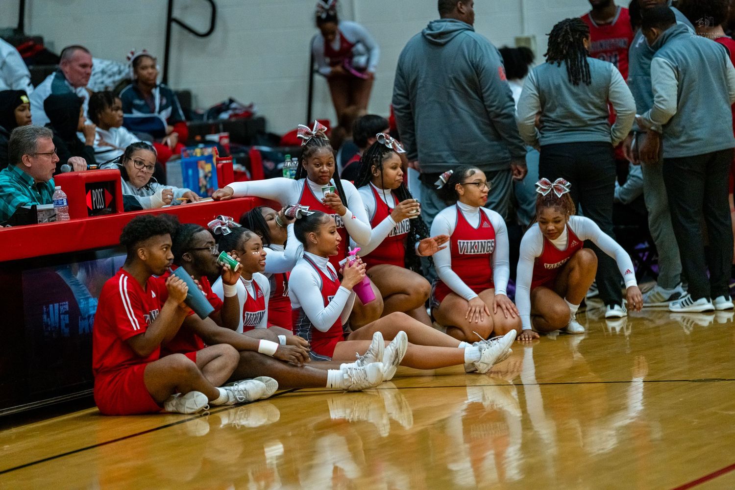 High school cheerleaders in red and white uniforms sit on the court floor during a basketball game.